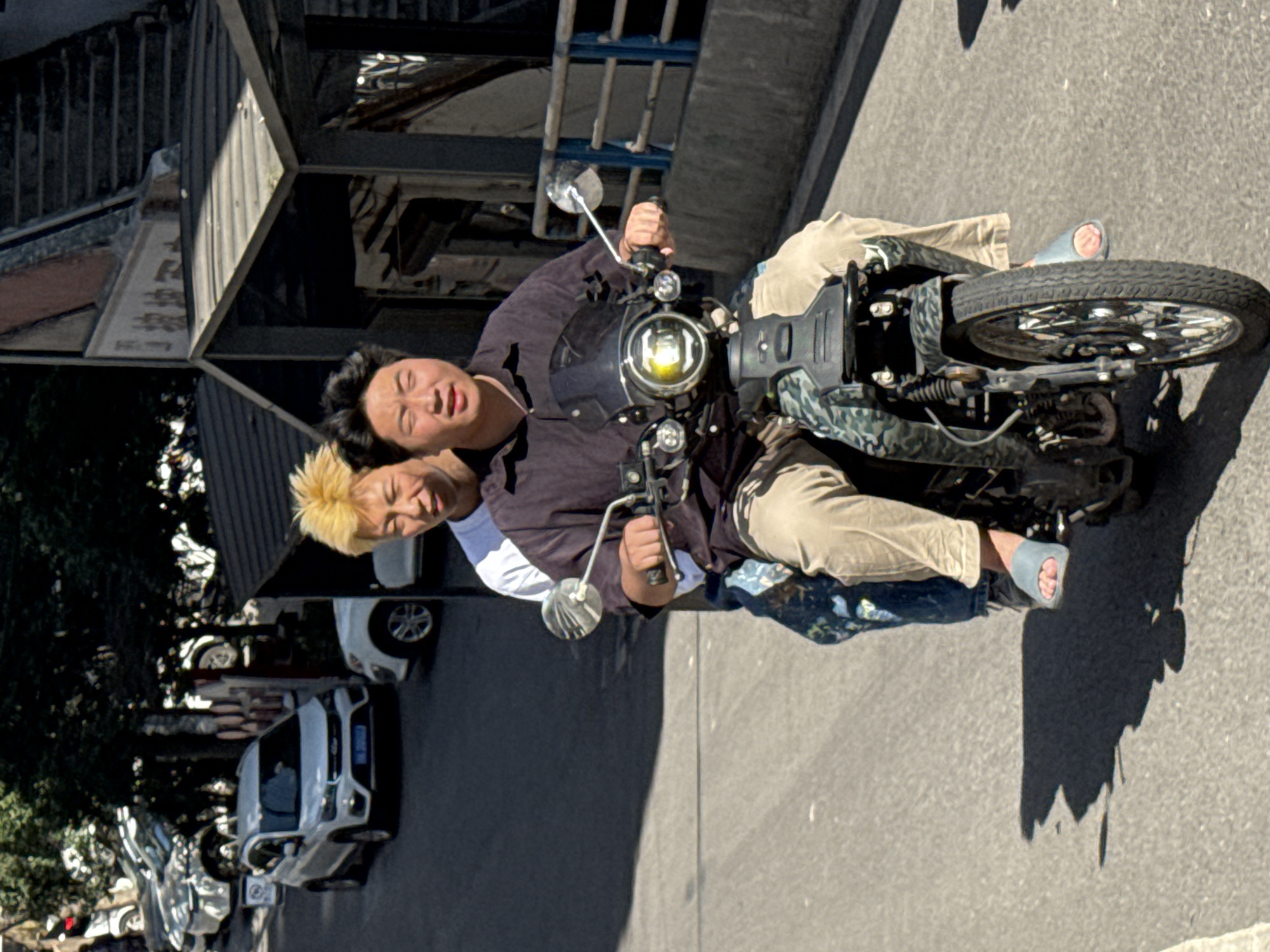 Two people are riding a motorcycle on a paved road in Jiulongpo District, China. The driver, with dark hair, wears a dark shirt and light pants. The passenger has blonde hair and wears a white shirt. The motorcycle has a round headlight and a camouflage pattern. Cars and trees are visible on the left side of the image, and buildings are in the background.