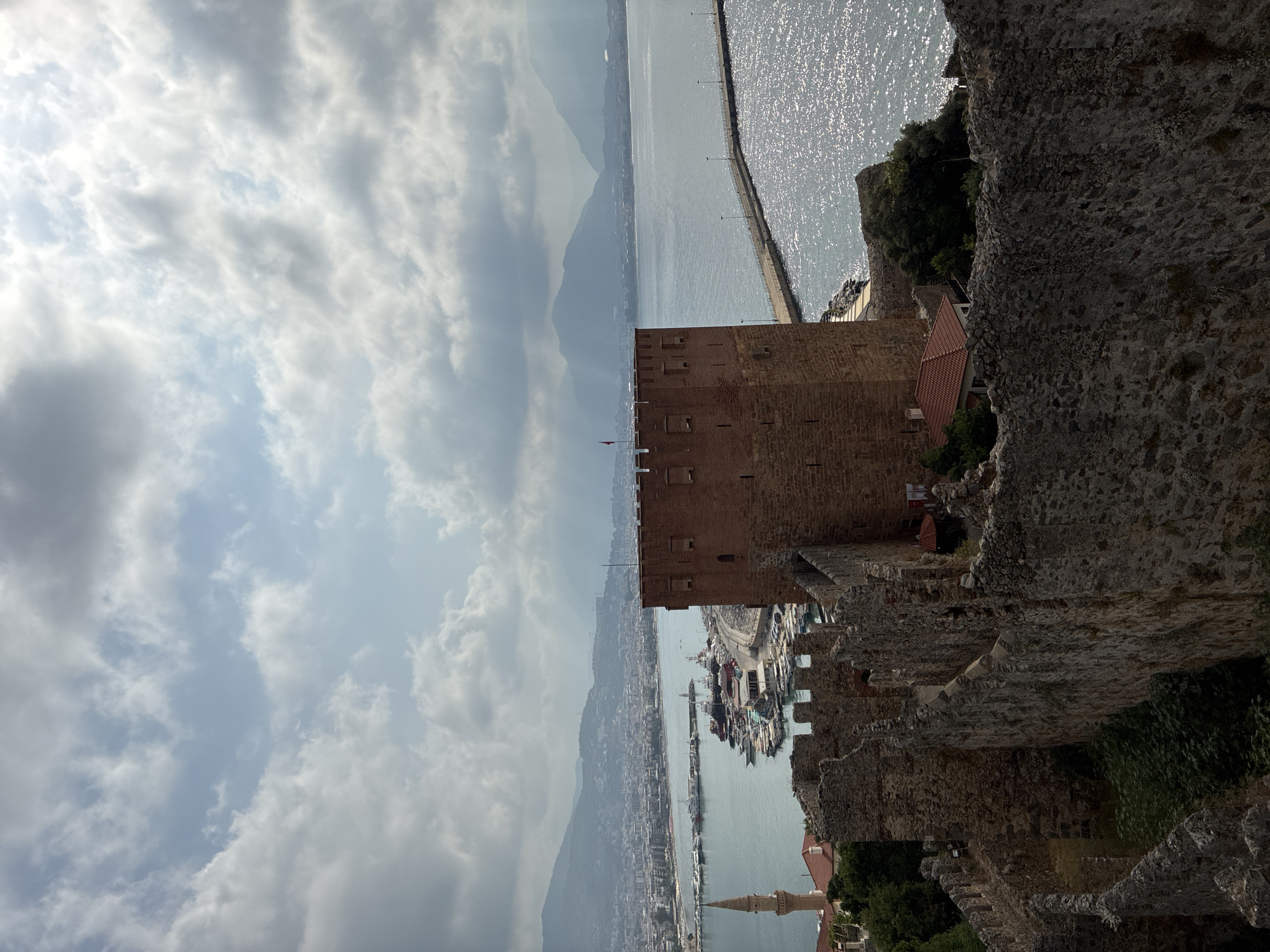 This is a high-angle, eye-level, medium shot of the Alanya Castle in Alanya, Turkey. The image is dominated by the castle walls and a prominent red brick tower. In the background, the blue sea stretches out, dotted with boats in a harbor and a long pier. A city with buildings and mountains can be seen in the distance under a cloudy sky. The foreground features the textured stone walls of the castle, with some greenery growing around them. The overall impression is one of historical architecture and a coastal landscape.