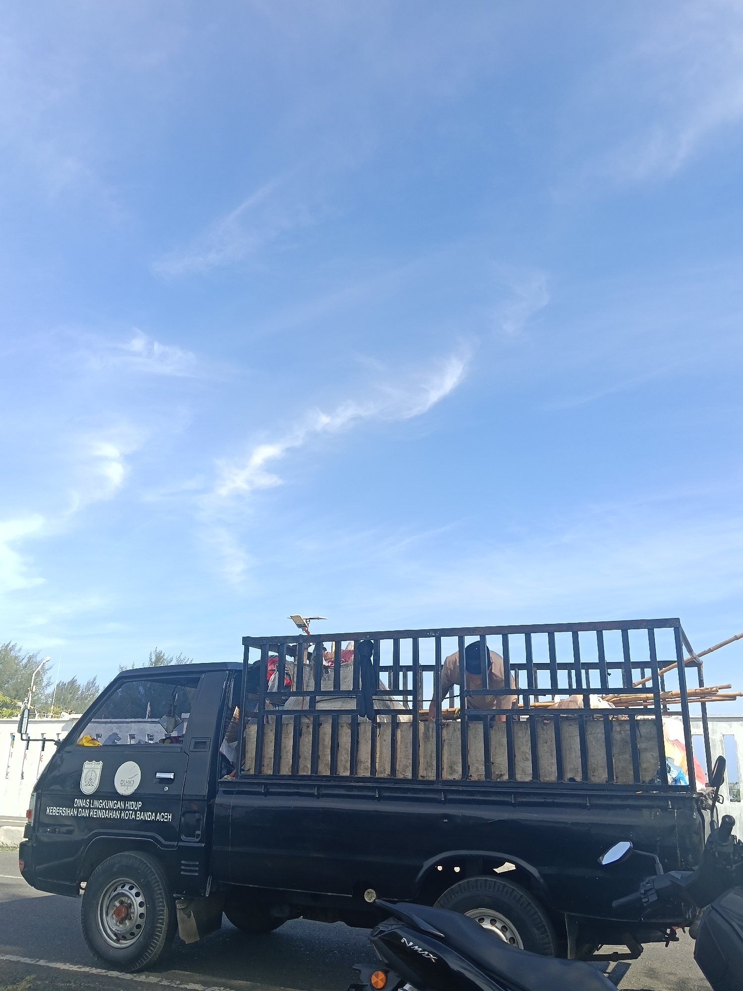 A black pickup truck is parked on a street in Banda Aceh, Indonesia. The truck is a Mitsubishi Colt T120SS. The side of the truck has text that reads "DINAS LINGKUNGAN HIDUP KEBERSIHAN DAN KEINDAHAN KOTA BANDA ACEH". The truck bed is filled with what appears to be building materials and possibly refuse, covered by a cage-like structure. A person wearing a brown shirt and a black cap is seen inside the truck bed. In the foreground, parts of two black motorcycles are visible. The sky above is clear blue with thin wispy clouds. Trees and some buildings are visible in the background on the left side of the image.