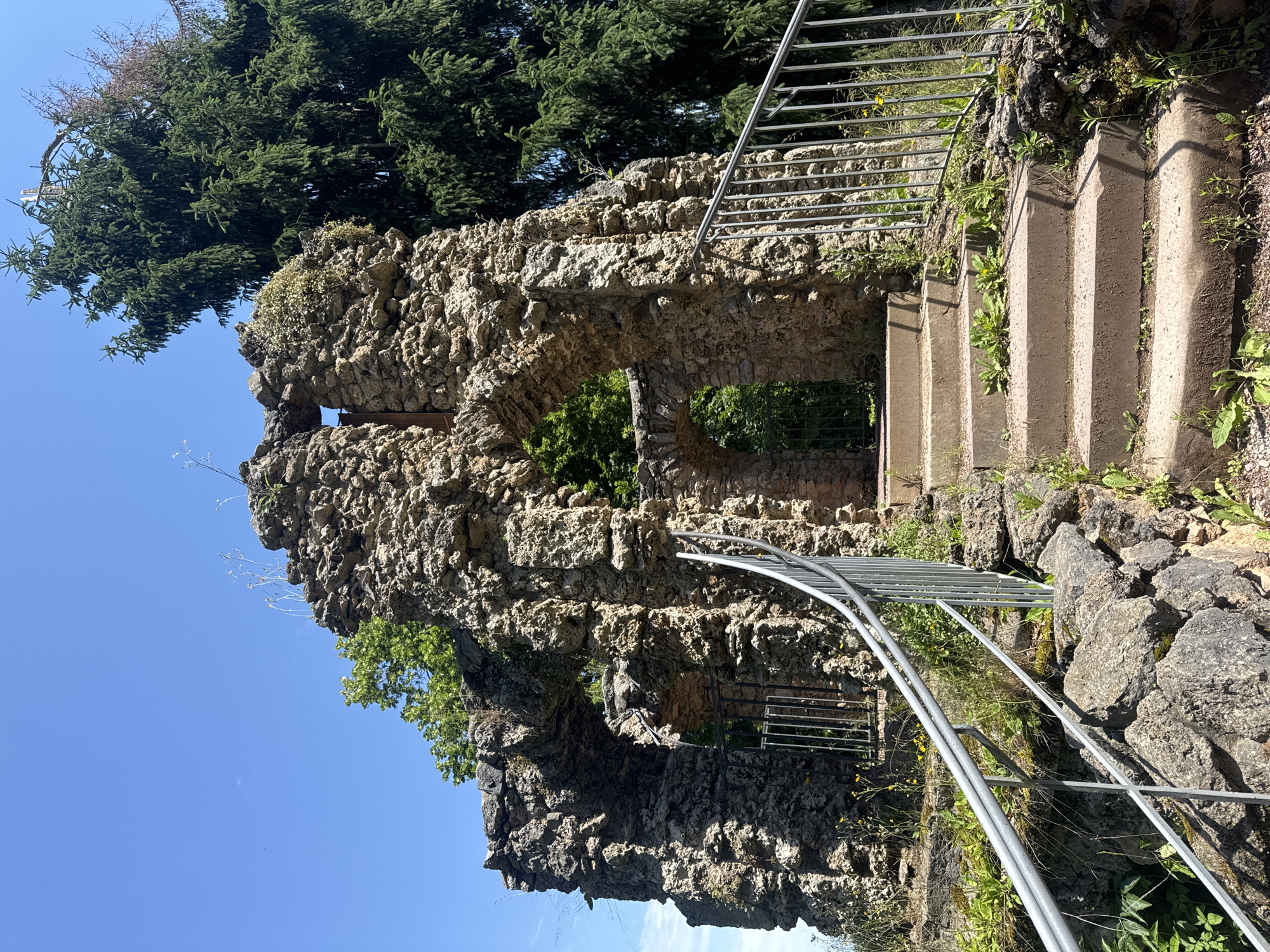 The image shows the Roman house ruins in Weimar, Germany, on a sunny day. A stone stairway with metal railings leads up to an arched entrance of the ruins. The ruins are made of rough, grey stones and have several archways and openings. Green trees, including a tall evergreen, are visible behind and around the ruins. The sky is clear blue. There are no people in the image.