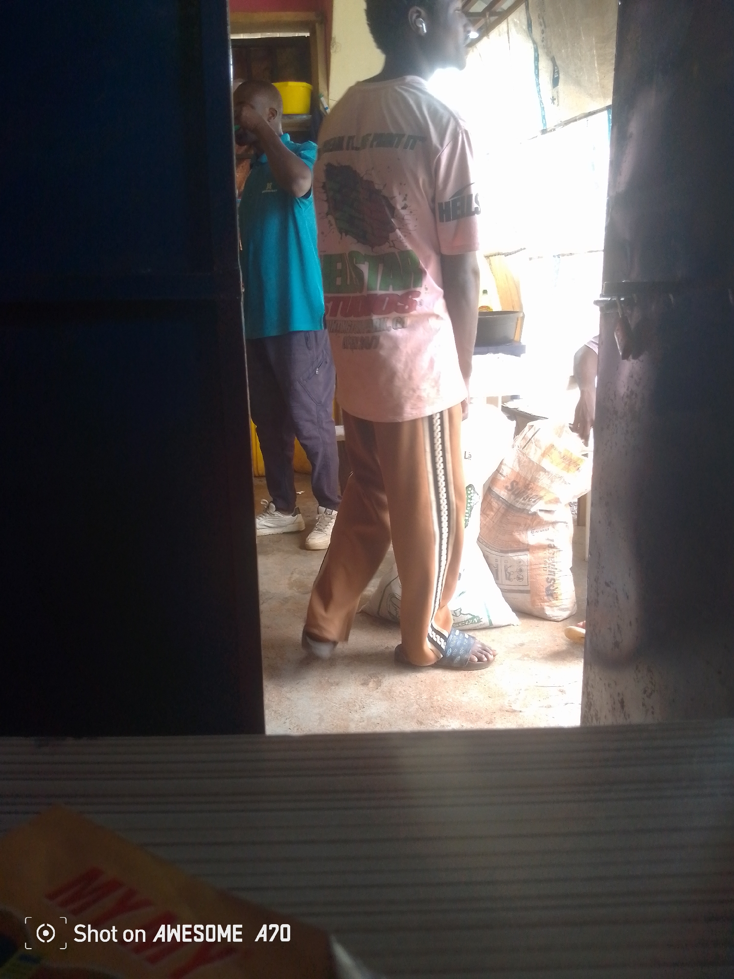 An outdoor scene features two men. One man, wearing a turquoise t-shirt and dark pants, stands on the left, holding a yellow bucket. The other man, in a pink t-shirt with graphics and brown pants, stands to the right, facing away from the camera. Bags of what appear to be supplies are piled behind him. The environment suggests a marketplace or storage area.