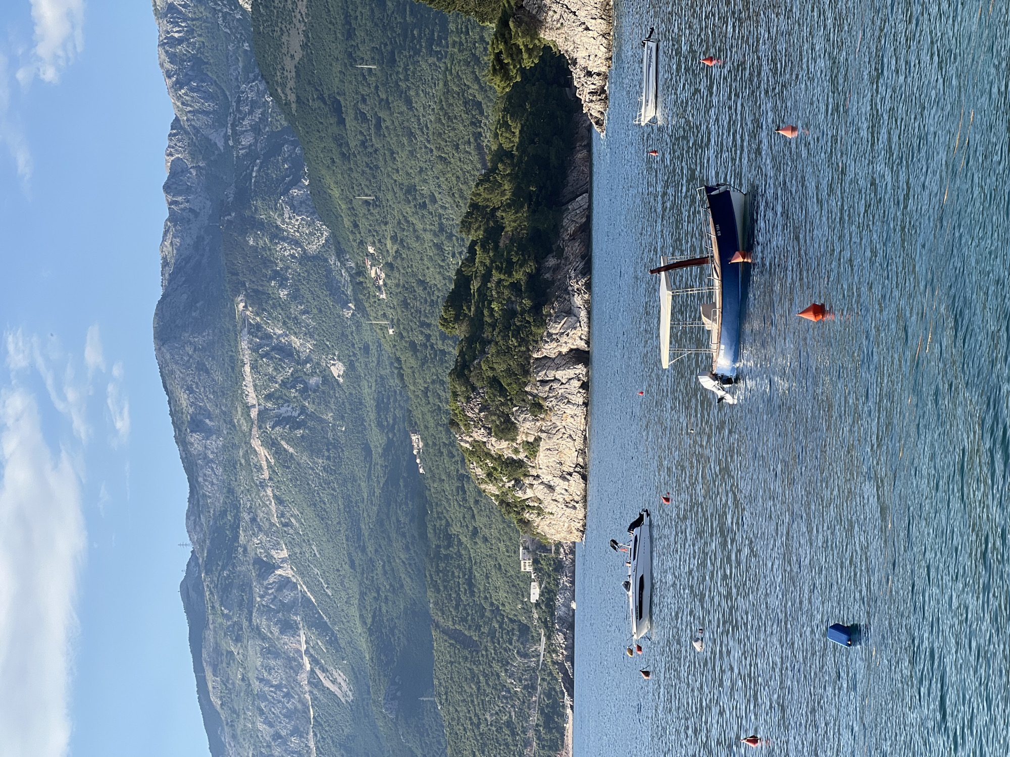 The image shows a coastal scene near Sveti Stefan, Montenegro. A tall mountain covered in green vegetation dominates the left side of the frame. The right side features a body of water, presumably the Adriatic Sea, with several boats moored offshore. Buoys are scattered in the water. Buildings are visible along the shoreline, nestled among the vegetation. The sky is clear with some scattered clouds.