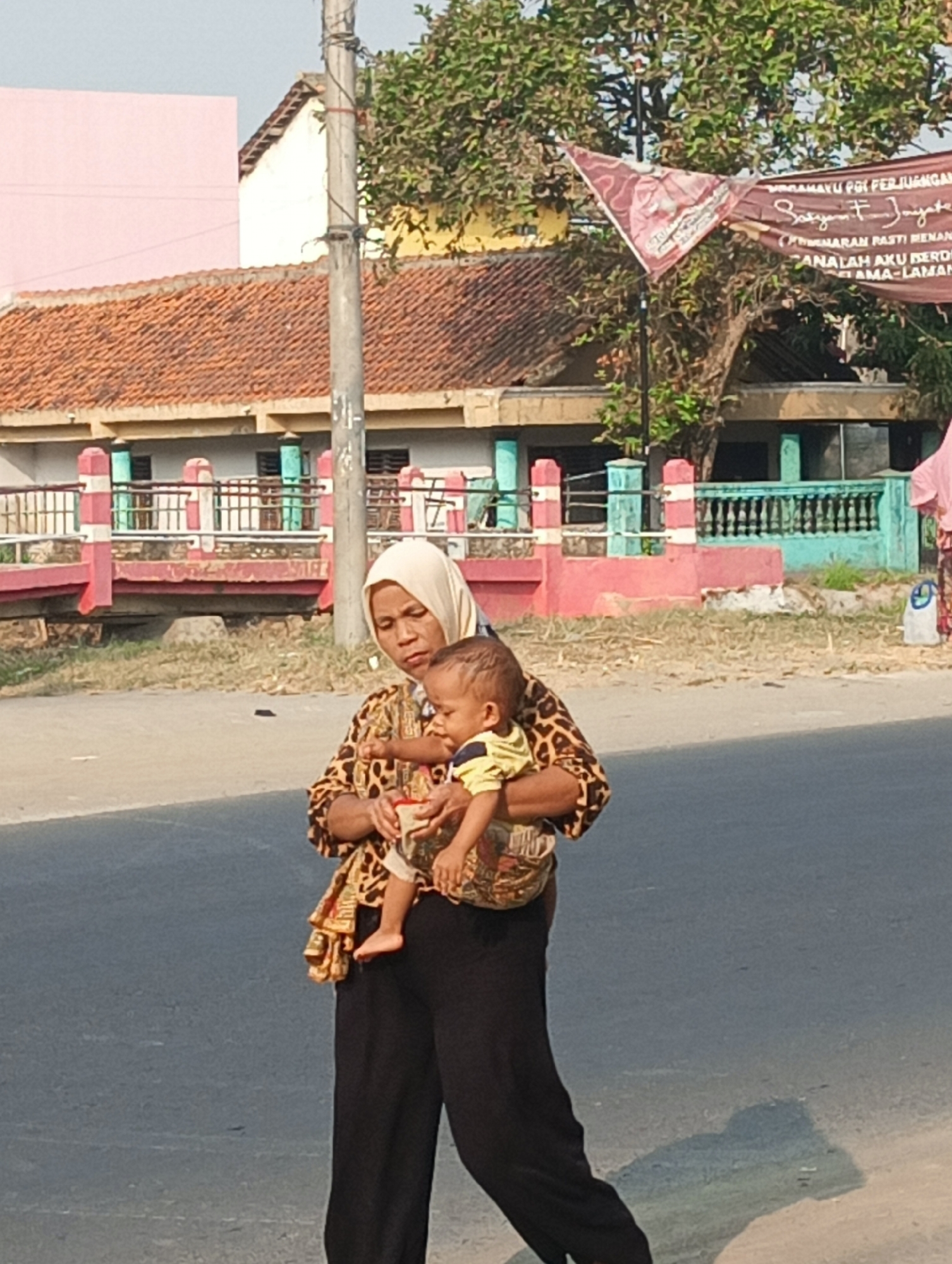 An Indonesian woman wearing a headscarf and a patterned top carries a baby wrapped in a fabric sling. She is walking on a paved road in front of a building with a red-tiled roof and a pastel-colored facade. The background includes a pole, trees, and a banner with Indonesian text. The scene appears to be in the city of Leuweunggajah, Indonesia.