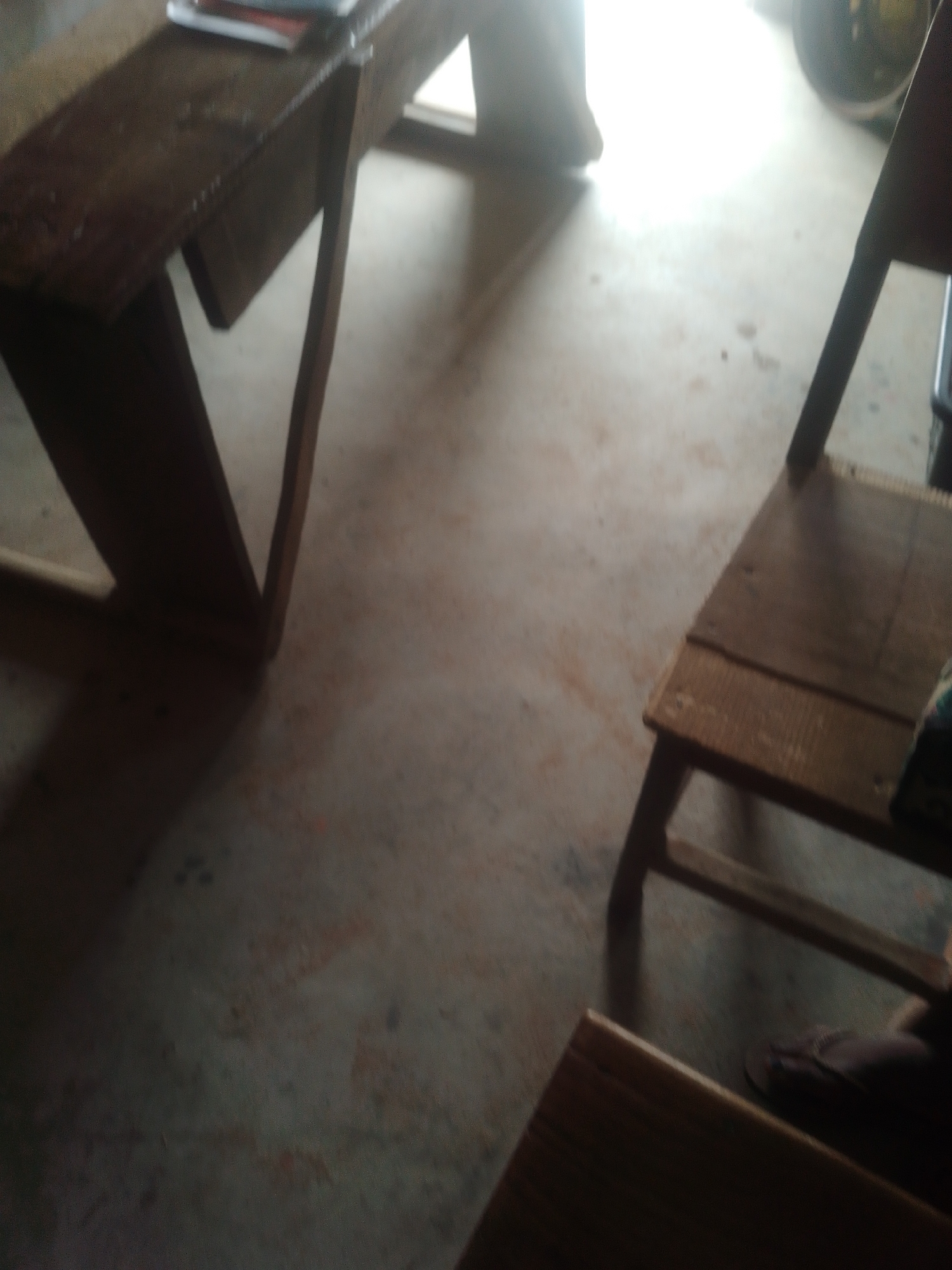 This image depicts an indoor scene, likely a classroom or a communal area, in Jalingo, Nigeria. The foreground is dominated by two wooden benches. The bench on the left is long and narrow with a tilted surface, suggesting it might be a desk or a reading bench. A stack of books or papers is visible on its surface. The bench on the right is shorter and appears to be a seating bench, with a person's feet, wearing dark flip-flops, visible at the bottom right corner. The floor is concrete, showing some dirt and reddish stains. The background is blurred and bright, indicating a light source, possibly a window or an open doorway, with a hint of a circular object, perhaps a fan or a wheel, to the far right. The overall ambiance is utilitarian and somewhat aged.