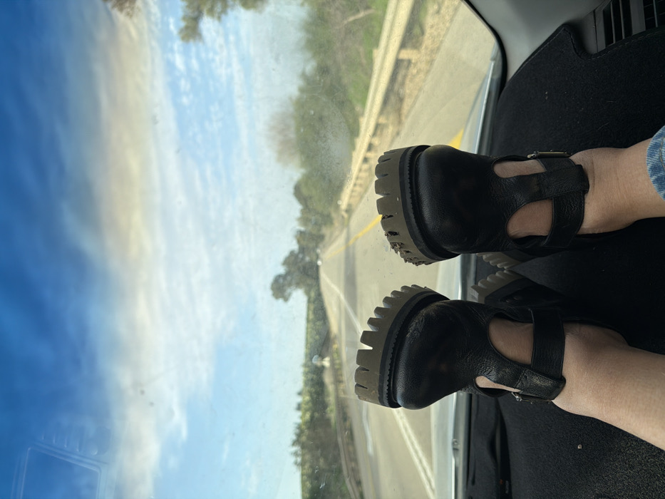 The image displays a first-person perspective from inside a vehicle, looking out over a road in the Hof Ashkelon Regional Council, Israel. Two feet are prominently propped on the black dashboard. These feet are clad in black, open-toed sandals with thick, serrated, dark-brown soles, one of which shows visible accumulated debris. The person's lower leg, partially visible on the right, wears light-wash denim. Through the vehicle's windshield, a two-lane asphalt road with a solid yellow center line extends into the distance, flanked by green vegetation and trees on both sides. A light-colored metal guardrail runs along the right side of the road. The sky overhead is partly cloudy with blue patches and scattered white clouds. A bridge-like structure is faintly discernible on the left side of the road in the mid-ground, and two small, distant vehicles are visible further down the road.