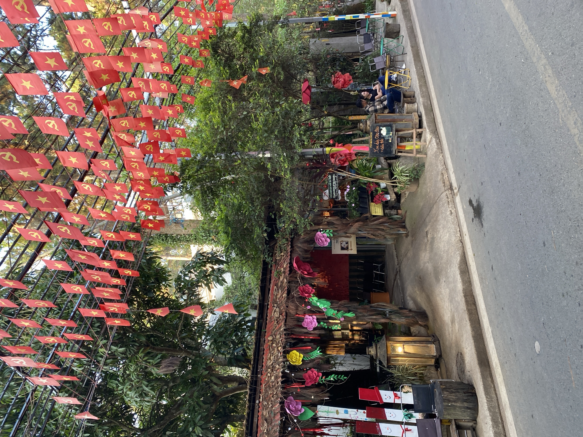 The image captures a street scene in Da Lat, Vietnam. Red flags with a yellow star and hammer/sickle are suspended overhead, creating a canopy. The flags are strung on a metal framework. Lush greenery, including trees and plants, is visible. A building decorated with paper flowers is present with a thatched roof. A person is sitting outside, near a blackboard menu. The road is to the right.