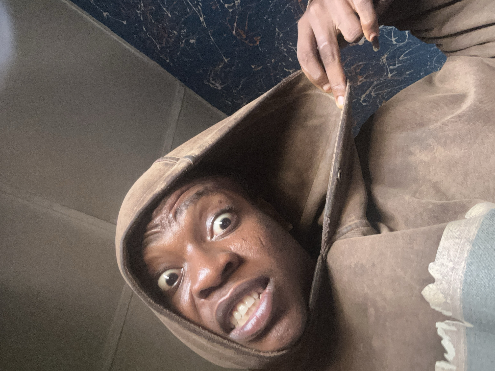 A close-up, tilted, low-angle shot depicts a dark-skinned person wearing a brown hooded sweatshirt. The person is pulling on the edge of the hood with their right hand. Their eyes are wide, and their mouth is open, revealing their teeth. The background appears to be a wall or ceiling with a dark pattern on it, suggesting an indoor setting. The location is Unknown, Unknown.