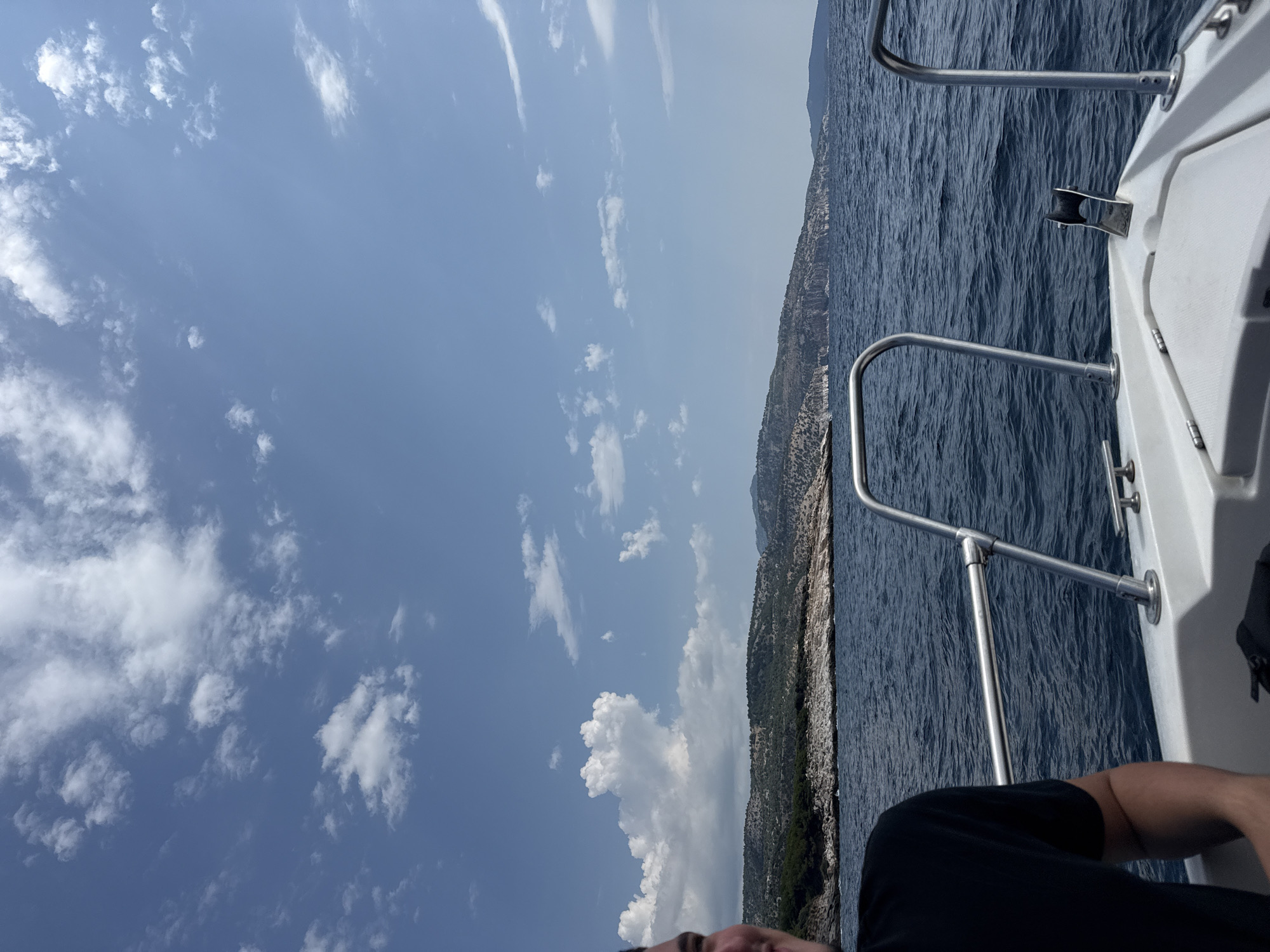 This is a vertical, outdoor, eye-level shot from a boat on the water, looking towards a rocky, tree-covered coastline under a partly cloudy blue sky. The foreground shows a white boat deck with silver railings and an anchor. To the left, a person wearing a black t-shirt is partially visible, looking to the left with a slight smile. The middle ground features the deep blue sea with small waves. The background displays a distant, hilly coastline with rugged terrain and scattered trees. The sky is a vibrant blue with scattered white, fluffy clouds. The overall scene depicts a sunny day at sea, likely in a coastal region of Greece, given the landscape features.