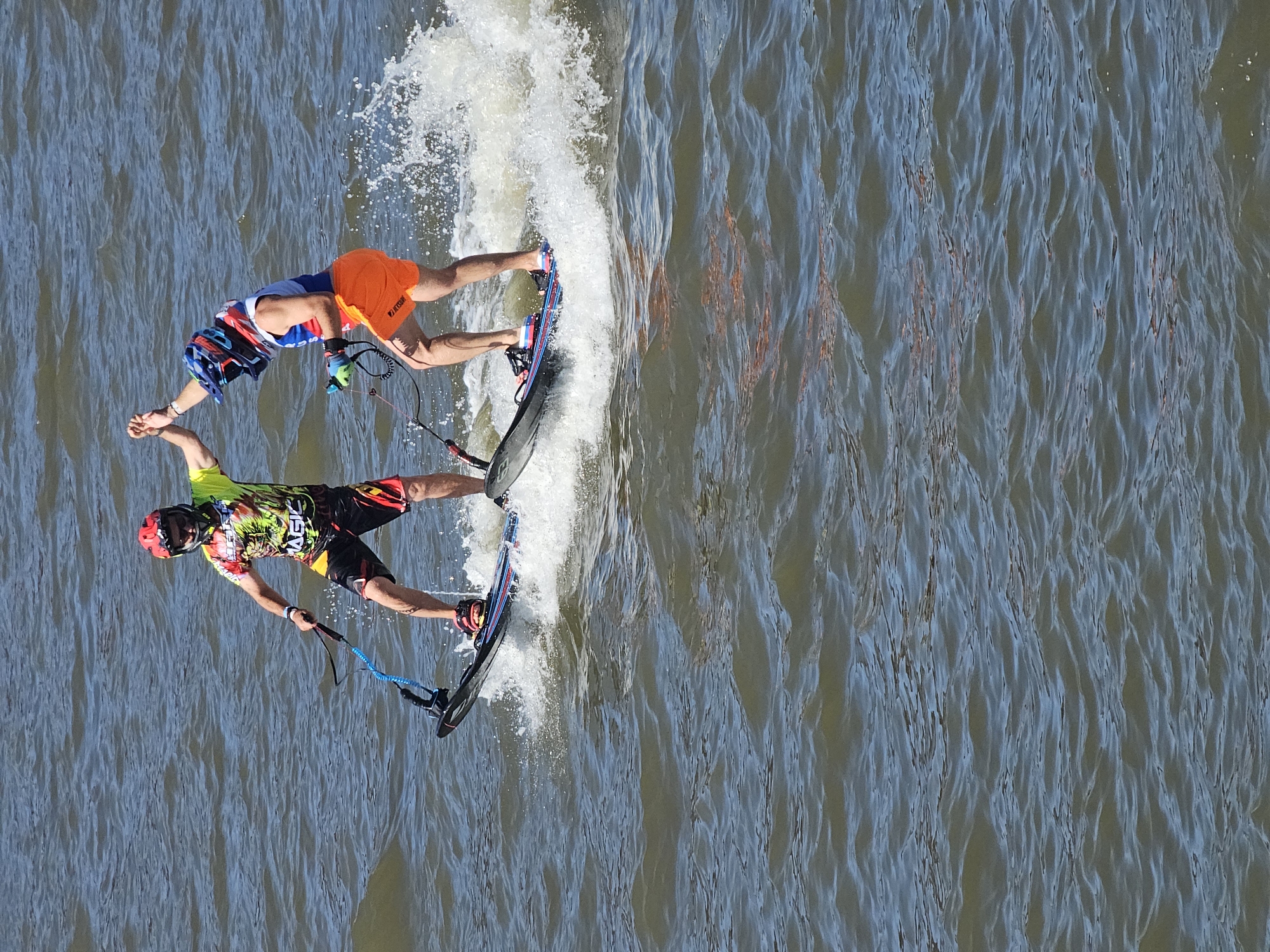 Two individuals are engaged in an aquatic activity, possibly jetboarding or a similar water sport, on a body of water. Both are wearing helmets, one red and the other a combination of blue and orange. The person on the left is wearing a brightly colored shirt with a graphic design and black and red shorts. They are holding onto a blue strap attached to their board. The person on the right is wearing a blue and white top with orange shorts and is also holding onto a strap connected to their board. The two individuals are high-fiving each other. They are standing on boards that are being propelled through the water, creating splashes around them. The water is choppy with visible ripples and waves. The background is composed of water, suggesting an outdoor, possibly open water environment. The lighting suggests it is daytime, with sunlight reflecting off the water.