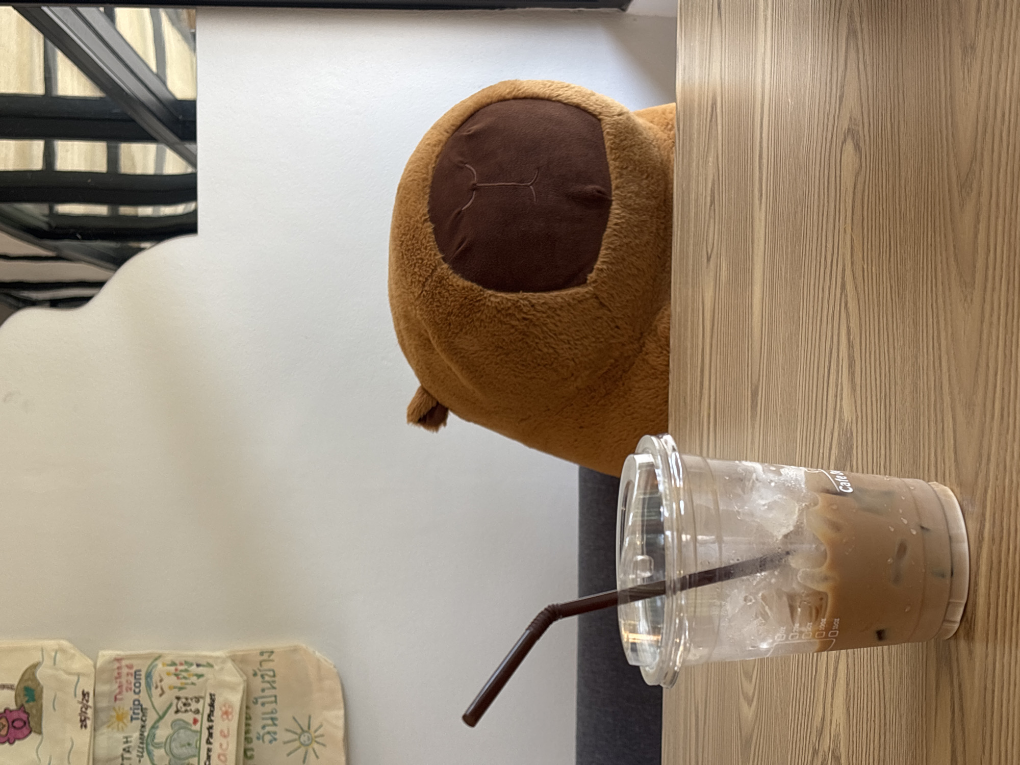 An indoor scene captures a partially consumed iced coffee drink next to a large, brown, plush object, likely a stuffed animal. The coffee, mostly liquid with some ice cubes remaining, is in a clear plastic cup with a brown straw, resting on a light-colored wooden table or surface. The plush object, which appears to be a large toy or pillow, is a fuzzy brown color with a darker brown, oval-shaped patch on its side, featuring a stitched design. This object is positioned behind the coffee cup, partially obscured by the table's edge.
The background features a white wall with a distinctive curved cutout on the left, revealing a dark, possibly metallic, railing structure behind it. Further to the left, at the bottom of the image, are several small, off-white fabric pouches or bags with colorful hand-drawn designs and text.
Visible text from the pouches includes "25/12/25" (a date, possibly in Thai Buddhist calendar), "TAH", "2026", "Trip.com", "Care Park Phuket", "Lace ge", and "ฉันเป็นช้าง" (Thai for "I am an elephant"). The side of the coffee cup also shows faint markings like "0100%" and "01502", and partially visible text "Cate", likely part of "Cafe". The setting suggests a casual environment, possibly a cafe or a lounge area in Choeng Thale, Thailand, given the location context and Thai script. The time of day is not discernible from the indoor lighting.