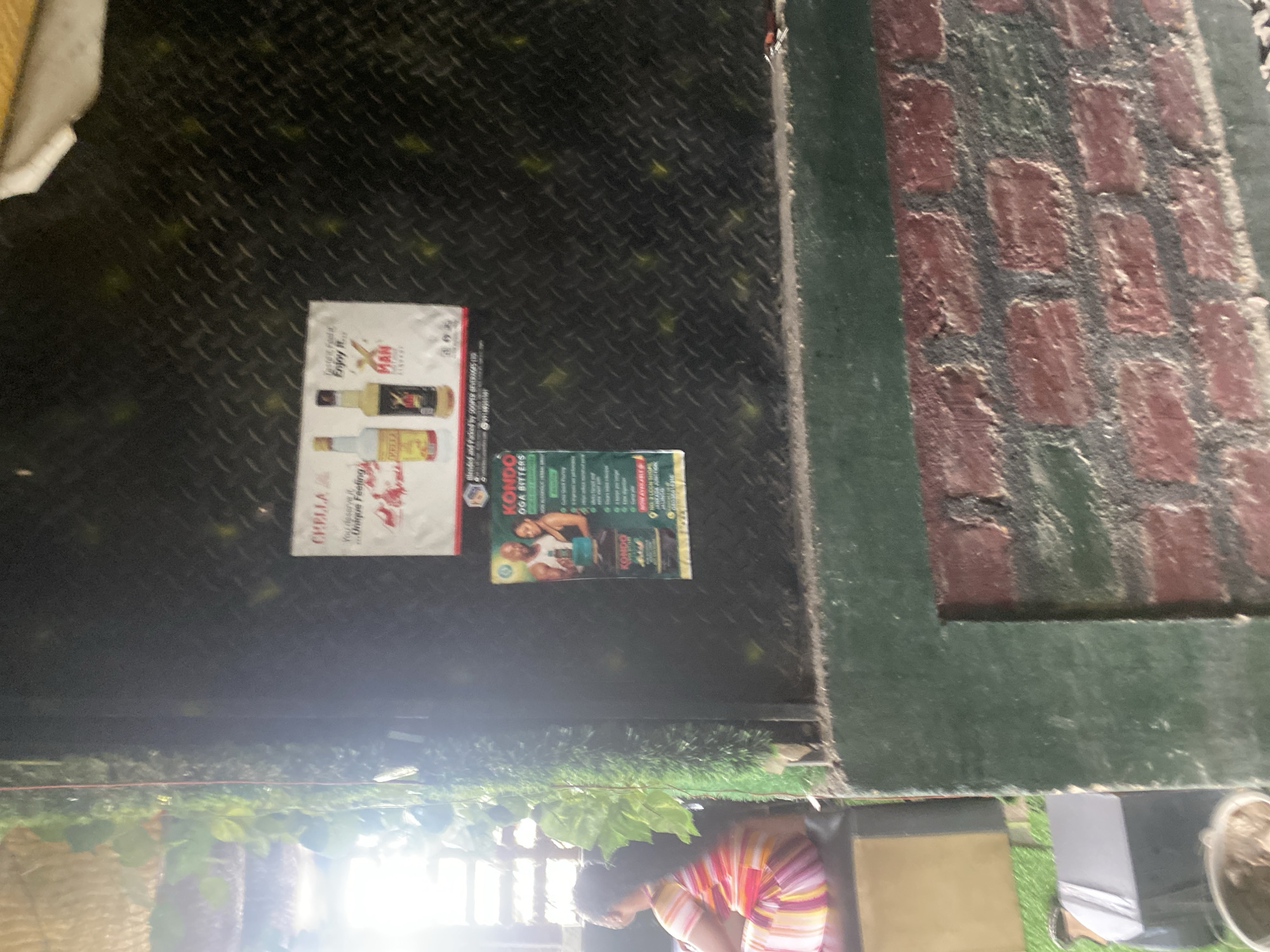 This image captures a scene with two posters displayed on a dark, textured surface, likely a wall or a counter. The poster on the left is primarily white with images of two bottles of what appears to be alcoholic beverages, one yellow and one clear. The text on this poster includes "CHELLA" and "Enjoy it...Unique Feeling". Below this, the text "MAN" is visible.
The second poster, positioned to the right of the first, is predominantly green and displays images of two men, one with a muscular physique. Text on this poster includes "KONDO" and "OGA BITTERS". There is also text indicating it is "NOW AVAILABLE" and mentions "JALINGG", which could be a location.
In the background, a brick wall with a dark green border is visible on the right side of the image. The lower portion of the image shows a glimpse of people and greenery, suggesting an outdoor or semi-outdoor setting. The lighting suggests it might be daytime, with some bright light sources in the lower right, possibly from a window or lamp. The overall impression is a casual display of advertisements in a public or commercial space.