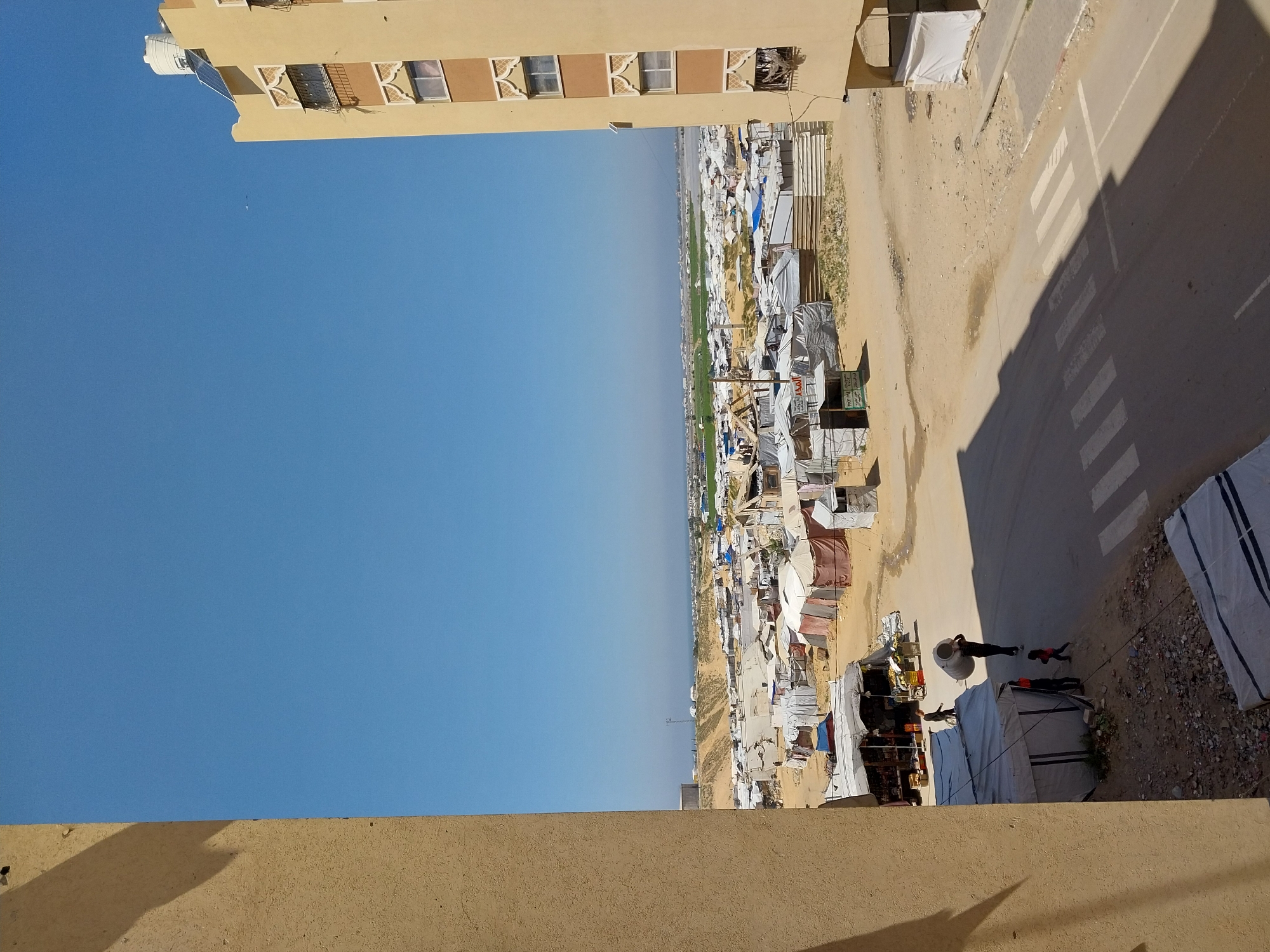 The image depicts an elevated view of a refugee camp adjacent to a paved road and a building, under a clear blue sky. The camp consists of numerous makeshift shelters constructed from tarpaulins, corrugated iron, and other materials. The paved road has a crosswalk with white stripes. Several people are visible near the road, one carrying a large container. The location is identified as the city of Unknown, Unknown.