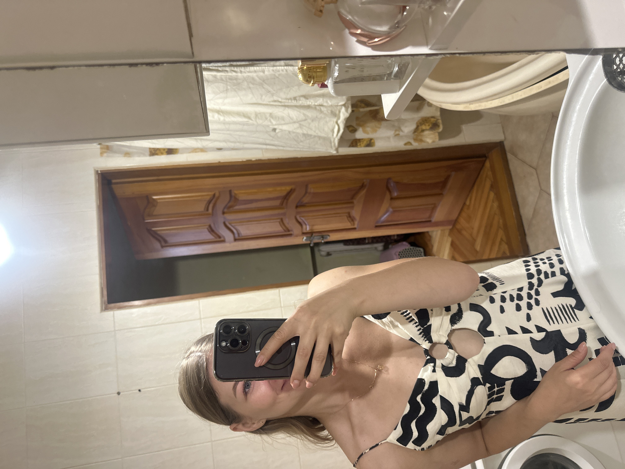 A young Caucasian woman with blonde hair pulled back is taking a selfie in a bathroom. She is wearing a black and white patterned dress with cutouts at the chest. She is holding a smartphone with a black case in her right hand, obscuring part of her face. In the background, a wooden door with carved panels is visible, as is a white tiled wall and a bathtub. A shelf with bottles of perfume is also present. The lighting suggests it is an indoor space. The scene appears to be a residential bathroom. The image does not contain any specific geographical markers or clues that would allow for definitive identification of the city as Ternopil, Ukraine.