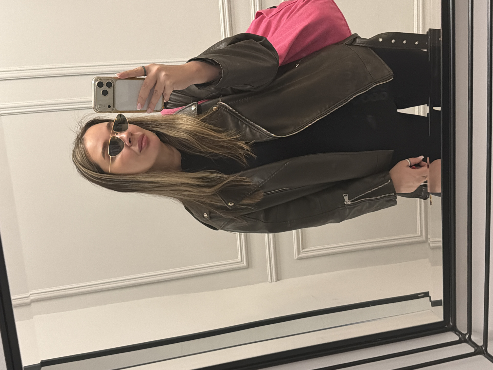A fair-skinned individual with long, light brown hair is taking a selfie in a mirror. They are wearing sunglasses, a black shirt, and a brown leather jacket. A pink shirt is visible underneath the jacket. The person is holding a smartphone in their left hand, and a ring is visible on their left ring finger. The background shows a white wall with decorative molding, and the mirror is framed in black. The location is not identifiable as Moscow, Russia.