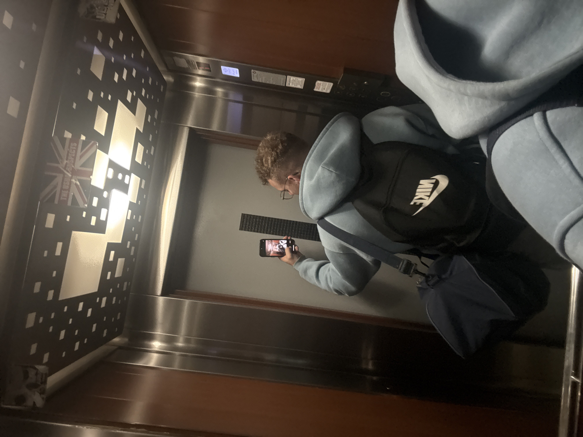 The image shows the interior of an elevator. A person is holding a smartphone, seemingly taking a picture of the elevator's interior. The person is wearing glasses, a light blue hoodie, and a black Nike backpack. A blue bag hangs from their shoulder. The elevator has a metal panel with a patterned design of squares and rectangles, and a stylized Union Jack flag is visible in the pattern. An elevator control panel is visible. Based on the design of the elevator, specifically the unique wall paneling with the Union Jack incorporated into the design, the location is likely the Ramada by Wyndham Plovdiv Trimontium Hotel in Plovdiv, Bulgaria.