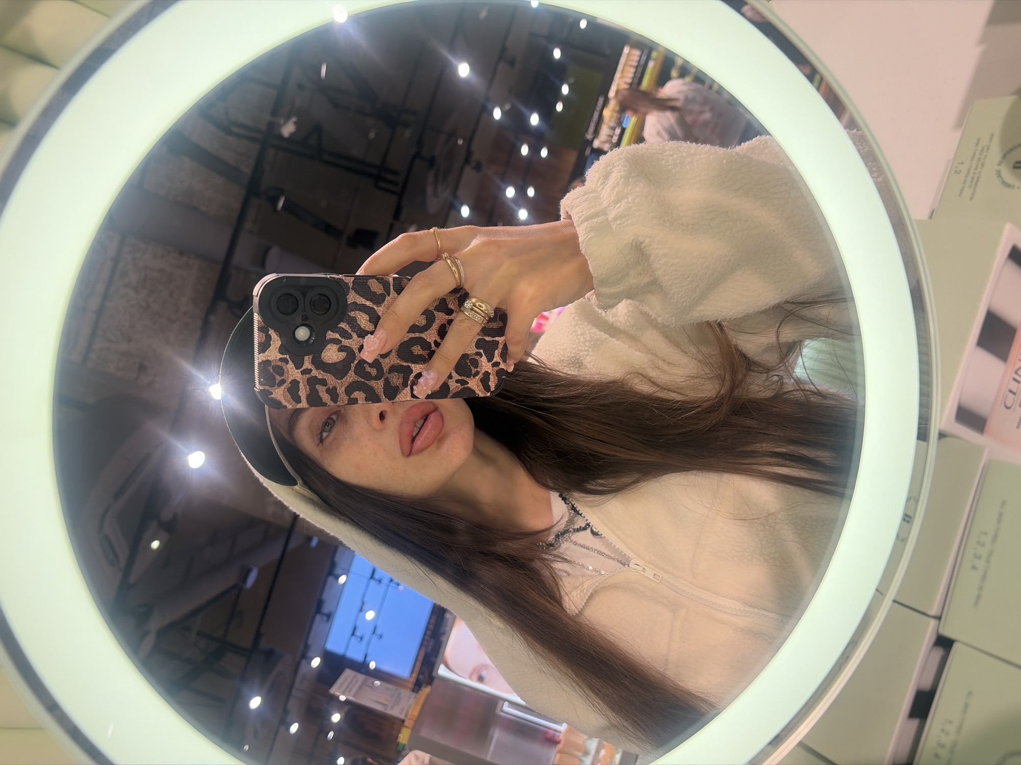 A young woman is taking a selfie in a circular mirror with a bright ring light. She is wearing a white fuzzy hooded jacket and a necklace. Her right hand is holding a leopard print phone case. She has long brown hair, light skin with freckles, and full lips. She is wearing rings on her fingers. The mirror reflects the interior of what appears to be a store, with shelves of products and overhead lighting. The scene suggests a casual, personal moment, likely in a makeup or beauty store given the products visible. The lighting is bright, indicating it is daytime, though the exact time of day is not specified. The image quality is good, with clear focus on the woman and her immediate surroundings.