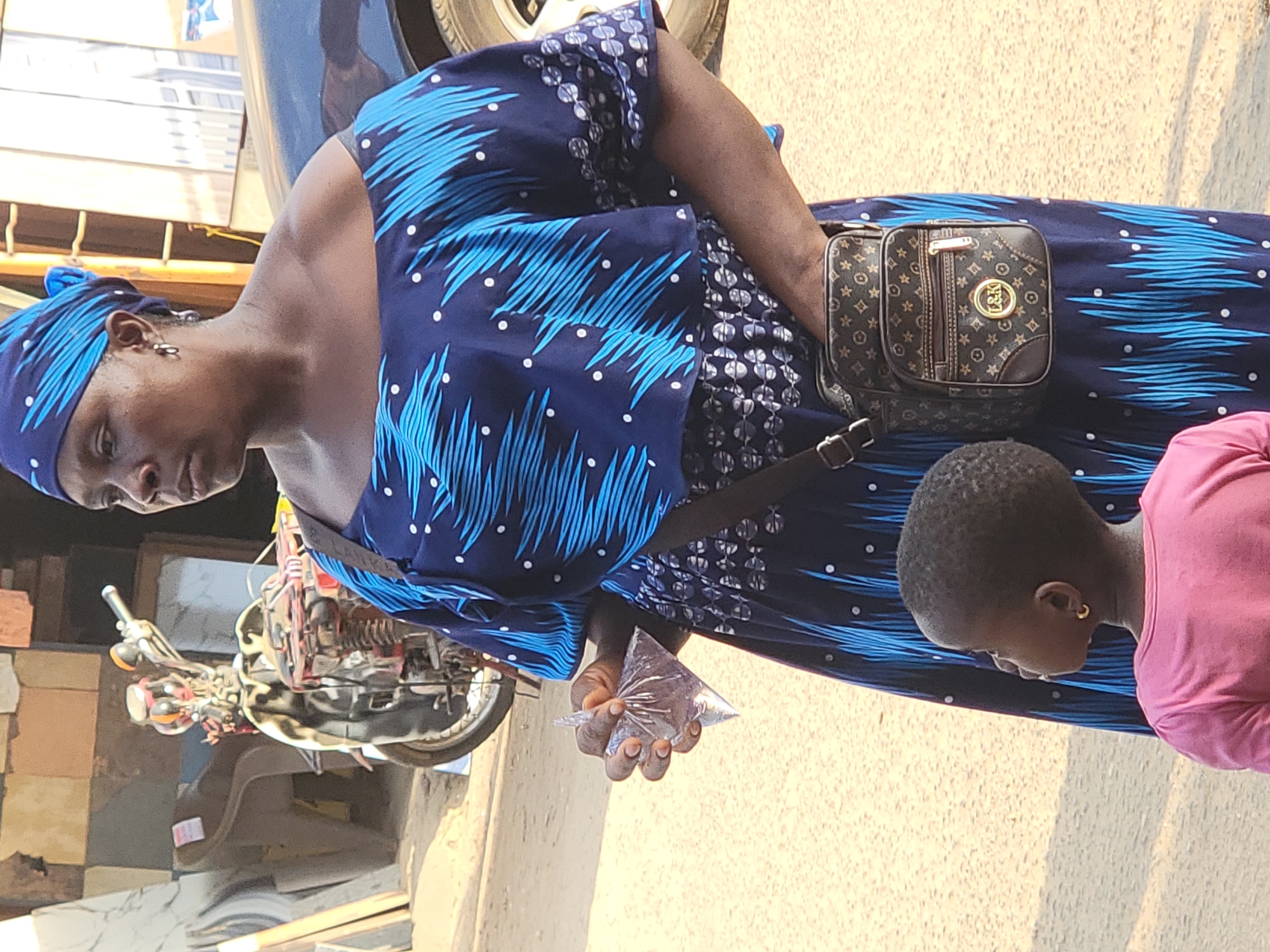 A medium shot captures an adult female wearing a blue patterned dress and matching headwrap in Maiburiji, Nigeria. She is holding a small transparent bag, possibly containing a liquid or small item. A child in a pink shirt stands adjacent to her. The woman has a small patterned purse slung across her body. In the background, a partially visible motorcycle sits on the side of the road, and the edge of a car's tire is visible above the woman's head. The setting appears to be an outdoor street scene.