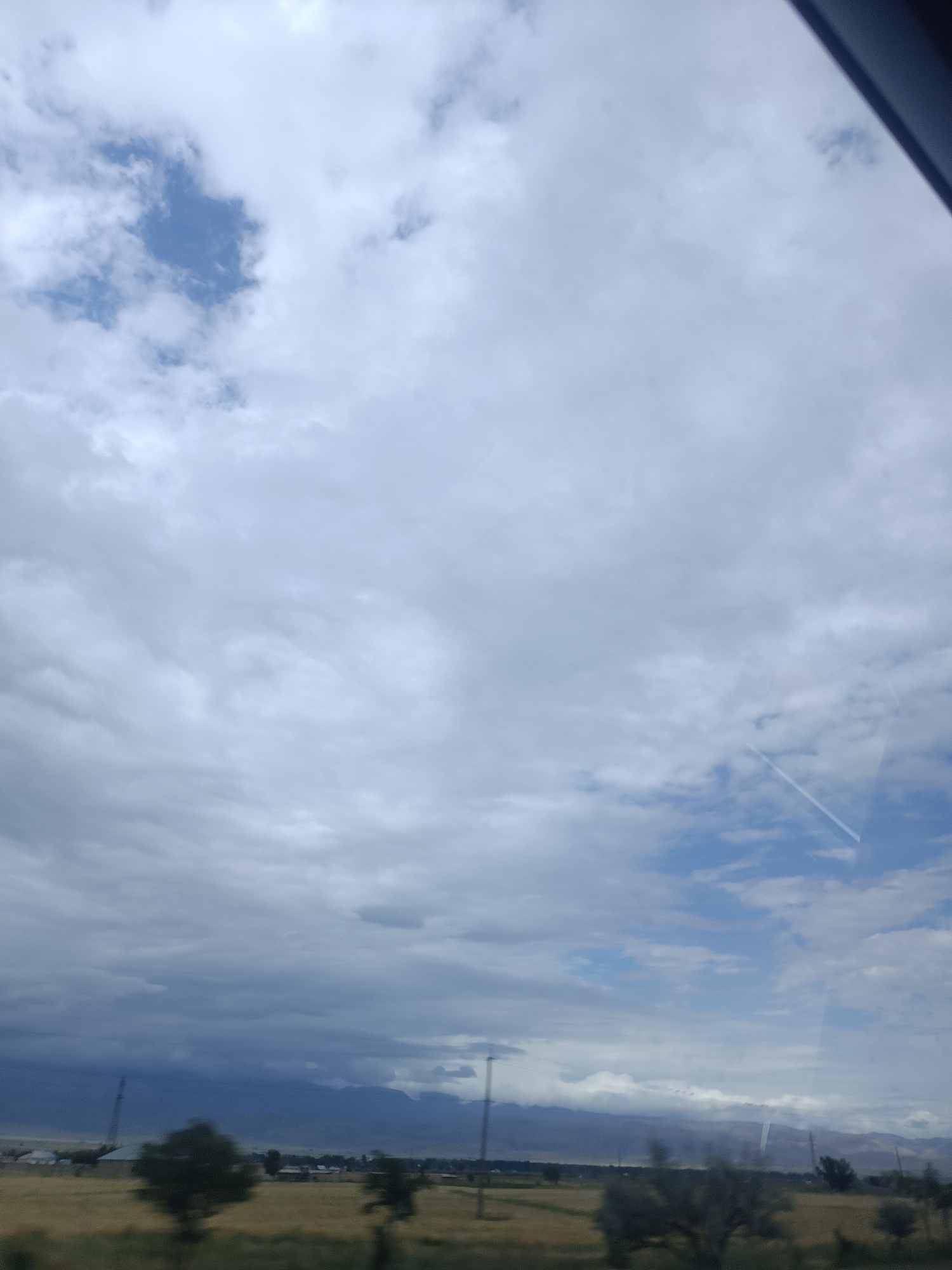 The image depicts a cloudy sky over a rural landscape, likely Sasyk-Bulak, Kyrgyzstan. The sky is filled with a mix of white and gray clouds, with patches of blue sky visible. A faint contrail from an aircraft is visible in the upper right portion of the sky. Below the clouds, a flat, dry field stretches towards a distant mountain range. Several sparse trees dot the landscape, and some low-lying buildings and utility poles are scattered throughout the scene. The overall impression is one of a somewhat overcast day in a rural, mountainous region.