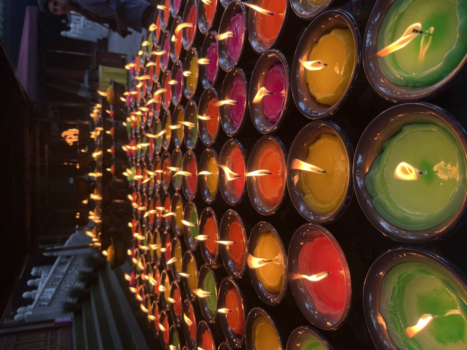 The image displays a structured arrangement of hundreds of lit candles within a low-light environment, located in the city of Shizhong, China. Each candle is contained within a small, shallow, reflective bowl. The candles exhibit various colors, including green, light green, red, orange, yellow, and magenta. They are organized in distinct rows and columns, extending from the foreground towards the background, with each candle showing an active flame. In the left background, a stone staircase with a white balustrade is visible, indicating a building or temple structure. In the right background, a male individual, partially obscured and out of focus, is present, viewed from the chest up. He wears a dark shirt and has a strap, possibly from a bag or camera, over his shoulder, and appears to be observing the scene. Additional blurred arrangements of lit candles on elevated surfaces are discernible in the distant background.