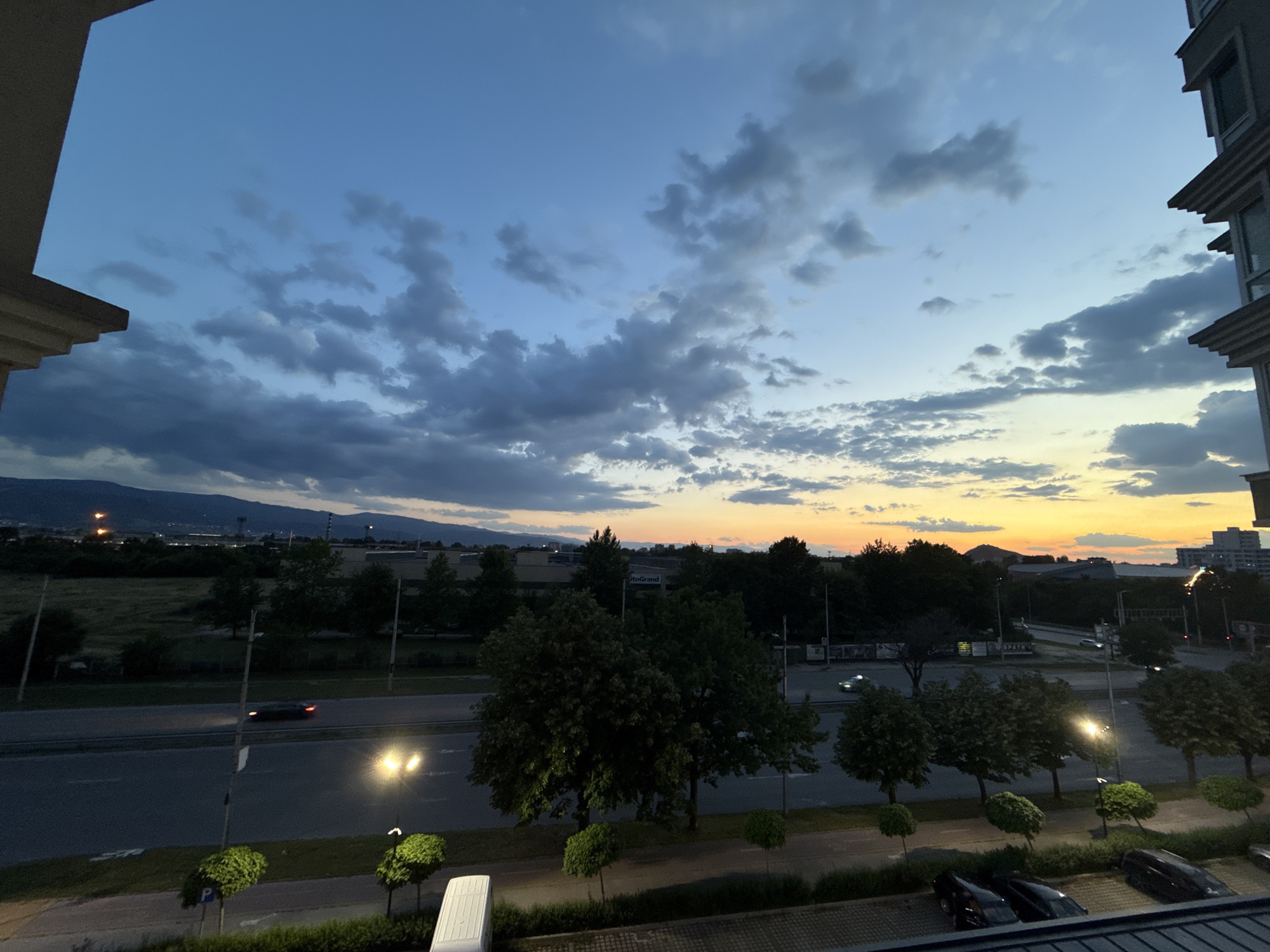The image shows an elevated view overlooking a road and landscape at dusk. The sky is a mix of blue and orange hues, with scattered clouds. A mountain range is visible on the horizon.
Below, a multi-lane road stretches across the scene, with cars traveling in both directions. Streetlights illuminate the road. On either side of the road are rows of trees and greenery. The foreground includes a parking area with several parked cars. Buildings are visible on either side of the frame, suggesting an urban or suburban environment.
The location is likely within the city of Plovdiv, Bulgaria, given the surrounding landscape and urban context. However, a precise address or landmark is not discernable from the image.