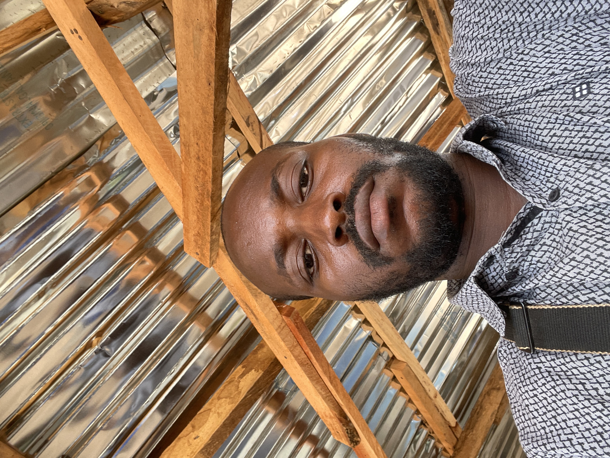 A medium shot captures a dark-skinned man with a shaved head and a full beard, wearing a patterned black and white button-down shirt and a black belt. The image is framed from a low angle, looking up at him, and it appears to be taken from indoors. The background consists of wooden beams and corrugated metal roofing. Based on the environment, the location may be in Angwan Sayawa, Nigeria.