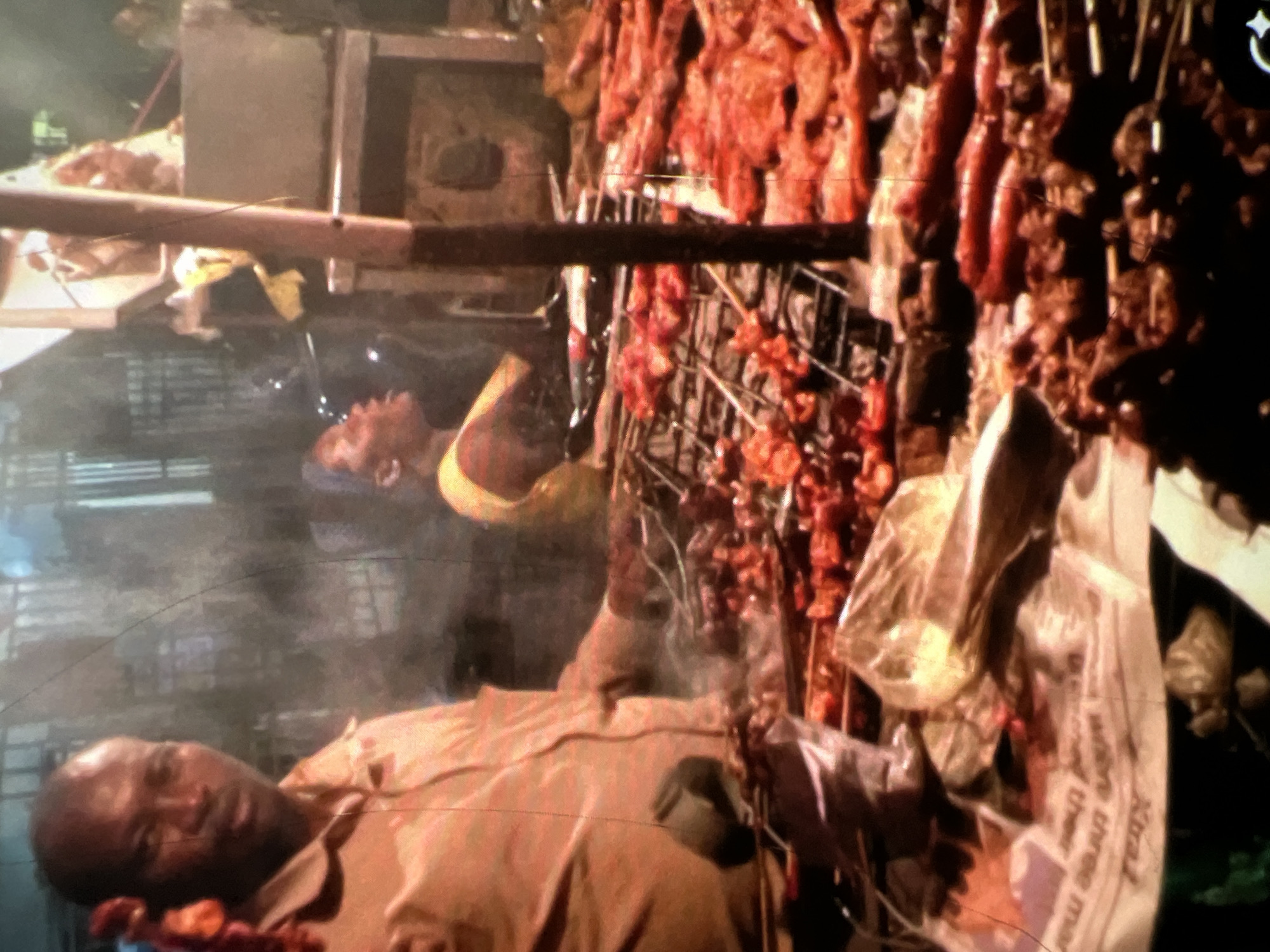 The image depicts a bustling night scene at a street food stall in Lagos, Nigeria, likely in a busy commercial or residential area where street vending is common. The central focus is a man, likely a vendor, wearing a tan collared shirt. He is positioned to the left, holding what appears to be meat on skewers, ready for grilling. In the background, another person, possibly a female customer, is partially visible wearing a yellow top and a headscarf, with her mouth open as if speaking or reacting.
The stall features a large grill laden with various meats, including what look like sausages and skewers of marinated meat, all in different stages of cooking. Smoke is rising from the grill, indicating active cooking. The foreground is partially obscured by newspapers, suggesting a makeshift setup for the vending operation, common in Lagos street markets. The overall atmosphere is lively and typical of a street food experience in the city, characterized by the aroma of grilled food and the din of activity. The lighting suggests it is night or dusk, illuminated by artificial lights from the stall and surrounding environment.