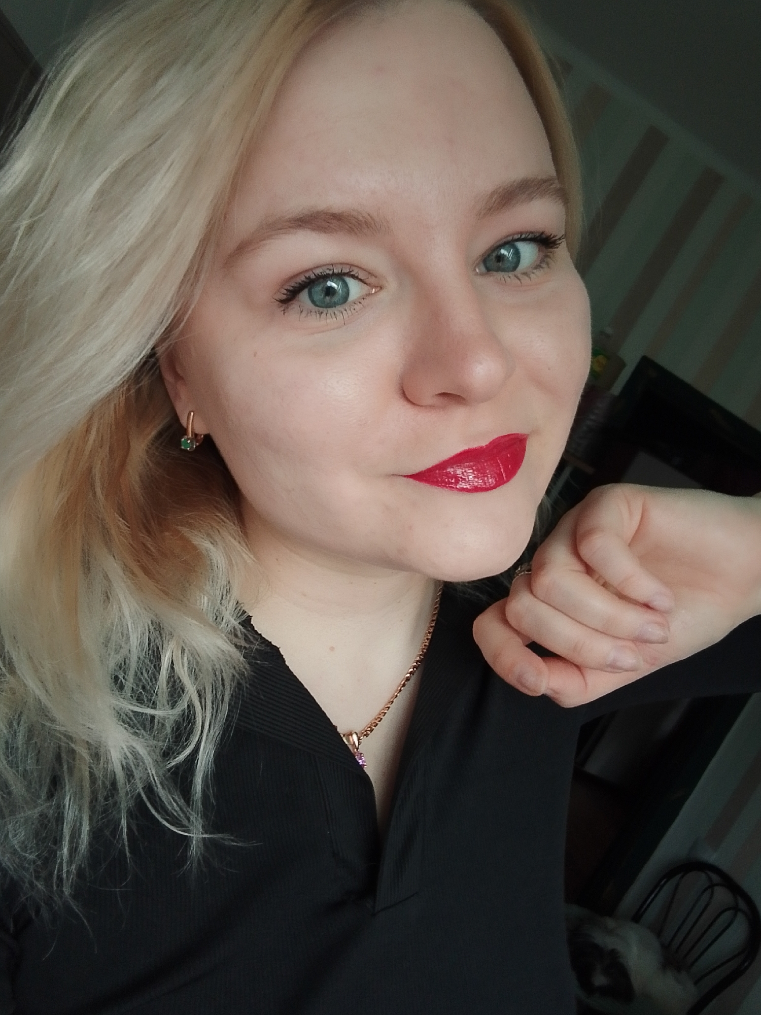 The image depicts an adult female in an indoor residential setting in Izhevsk, Russia. The subject has fair skin, blonde hair, and blue eyes, and is wearing red lipstick. She is dressed in a black long-sleeved top featuring a v-neck and ribbed fabric on the shoulders. Her accessories include a gold-colored chain necklace with a small purple pendant and gold-colored hoop earrings with green stones in her left ear. A simple ring is present on the ring finger of her left hand, which is held with fingers loosely curled near her chin. She looks directly forward with a subtle smile. The background includes a wall with vertical light and dark stripes, a dark framed object, a dark chair with a curved back, and a partially visible small, light and dark-colored animal resting near the bottom right.