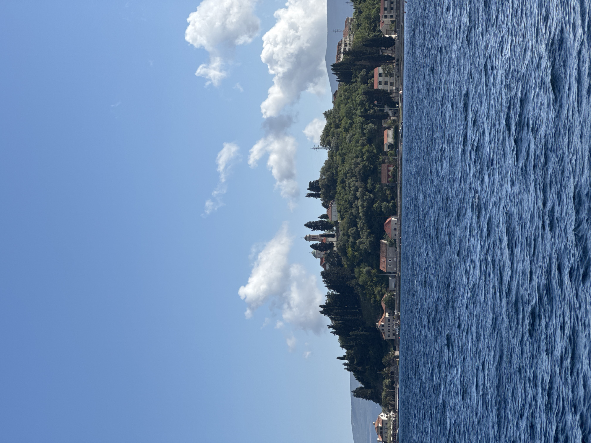 The image depicts a coastal view in Đurići, Montenegro. The sky is predominantly blue with scattered white clouds. A strip of land runs diagonally across the image, featuring dense green vegetation interspersed with buildings, including houses with red roofs and a church with a prominent dome. To the right of the land is a body of water, possibly the Adriatic Sea, with a textured surface reflecting the light.
