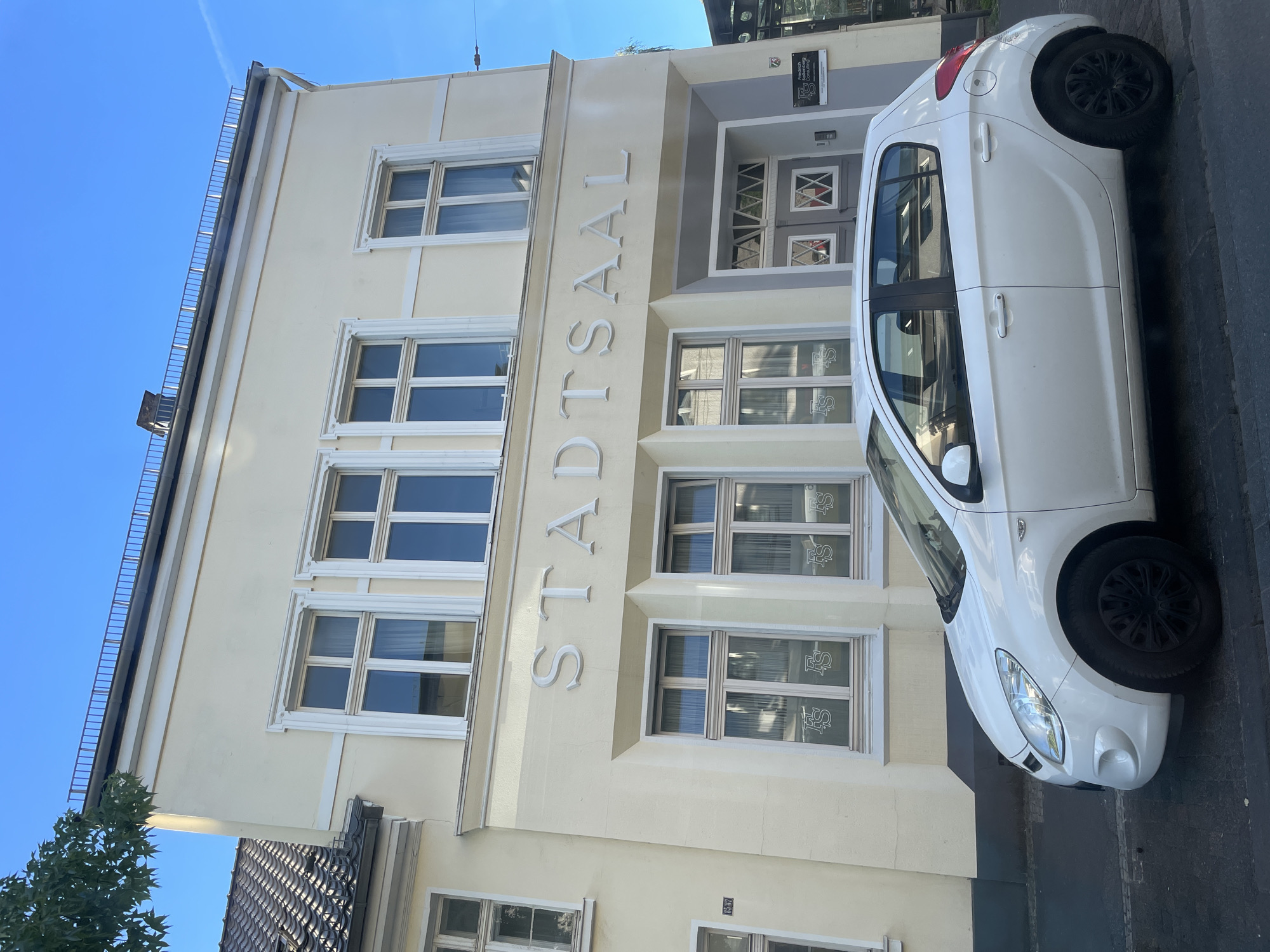 The image shows a white Hyundai i20 parked on a street in front of a building with the text "STADTSAAL" on its facade, indicating it is a city hall or a town hall. The building is a light yellow color with white trim around the windows. The windows are large and appear to be multiple panes. Above the "STADTSAAL" inscription, there are a row of four windows. Below the inscription, there is an entrance with a double door and a patterned glass transom. To the right of the entrance, a small sign is visible. The car is parked parallel to the curb. The sky is blue with some wispy clouds. The scene appears to be in Solingen, Germany.