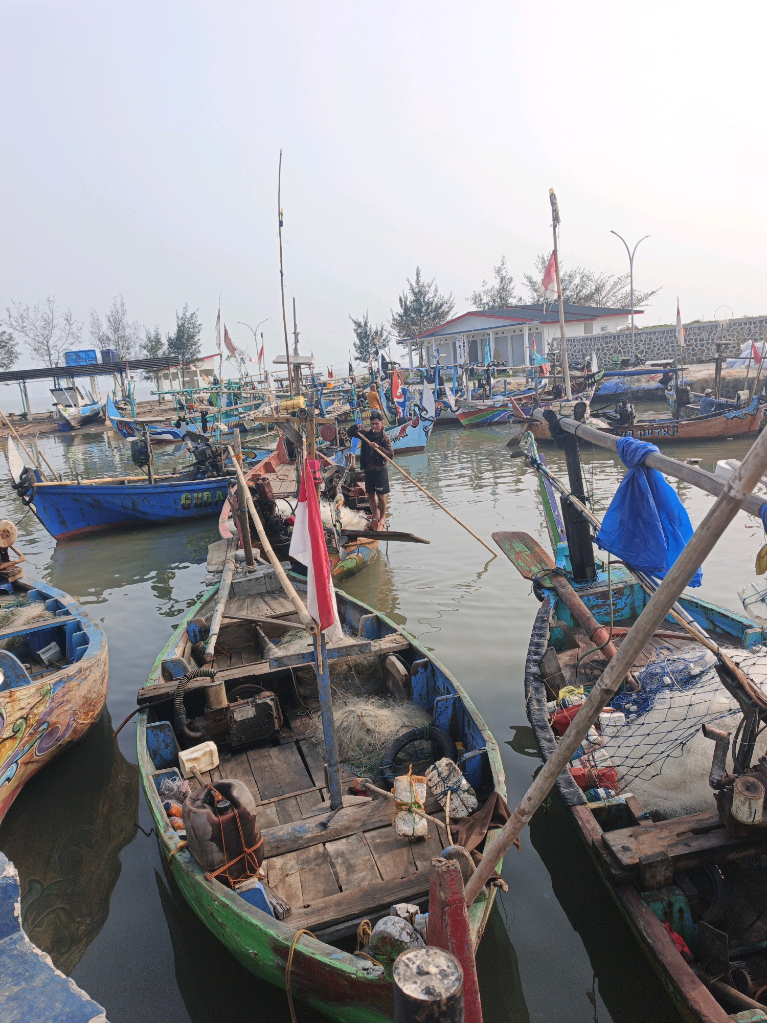 This is a photograph of a harbor in Bumiharjo, Indonesia, filled with numerous fishing boats. The boats are mostly wooden and appear to be of a traditional design, with various colorful decorations on some of them. Many of the boats are tied together or docked alongside each other. Fishing nets, paddles, and outboard motors are visible on and around the boats, suggesting their active use. In the foreground, a young person is standing in one of the boats, holding a long pole, possibly steering or preparing to push off. Another person is visible in a boat in the background. Indonesian flags are prominently displayed on several boats. The water in the harbor is calm and reflects the surrounding boats and sky. In the background, there are buildings with tiled roofs and a stone wall, and some trees can be seen. The sky is overcast, suggesting a hazy or cloudy day. The overall scene depicts a working fishing harbor.