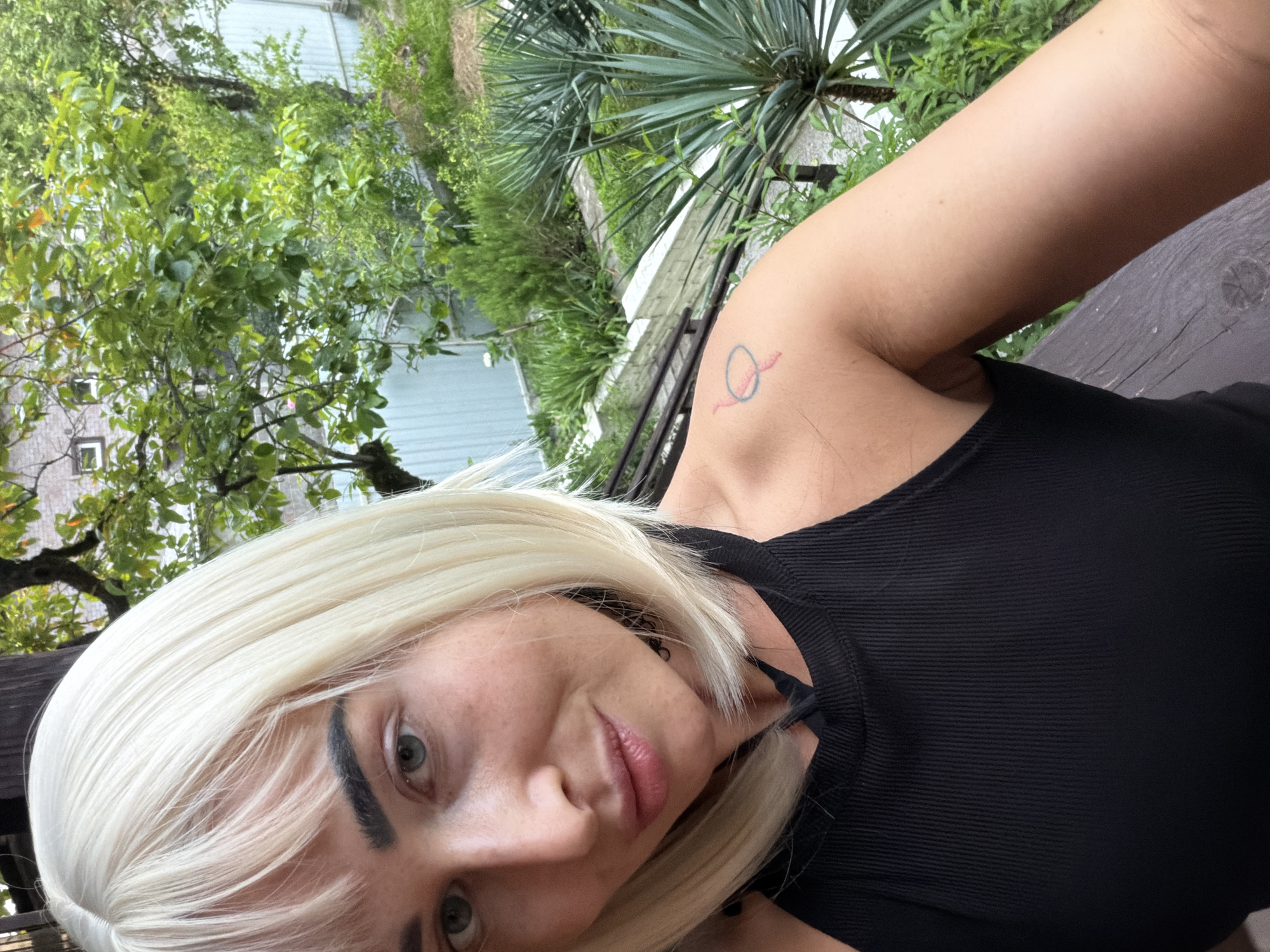 A close-up selfie of a woman with blonde hair and bangs is taken outdoors. She is wearing a black ribbed tank top. A small, abstract tattoo of a circle with a line through it and two small wing-like shapes is visible on her left shoulder. The background shows lush greenery, including trees and plants, and a light blue corrugated metal fence. The scene appears to be in a garden or yard setting, possibly on a balcony or patio, given the wooden railing in the foreground. The image is likely from Sochi, Russia, based on the presence of subtropical vegetation.