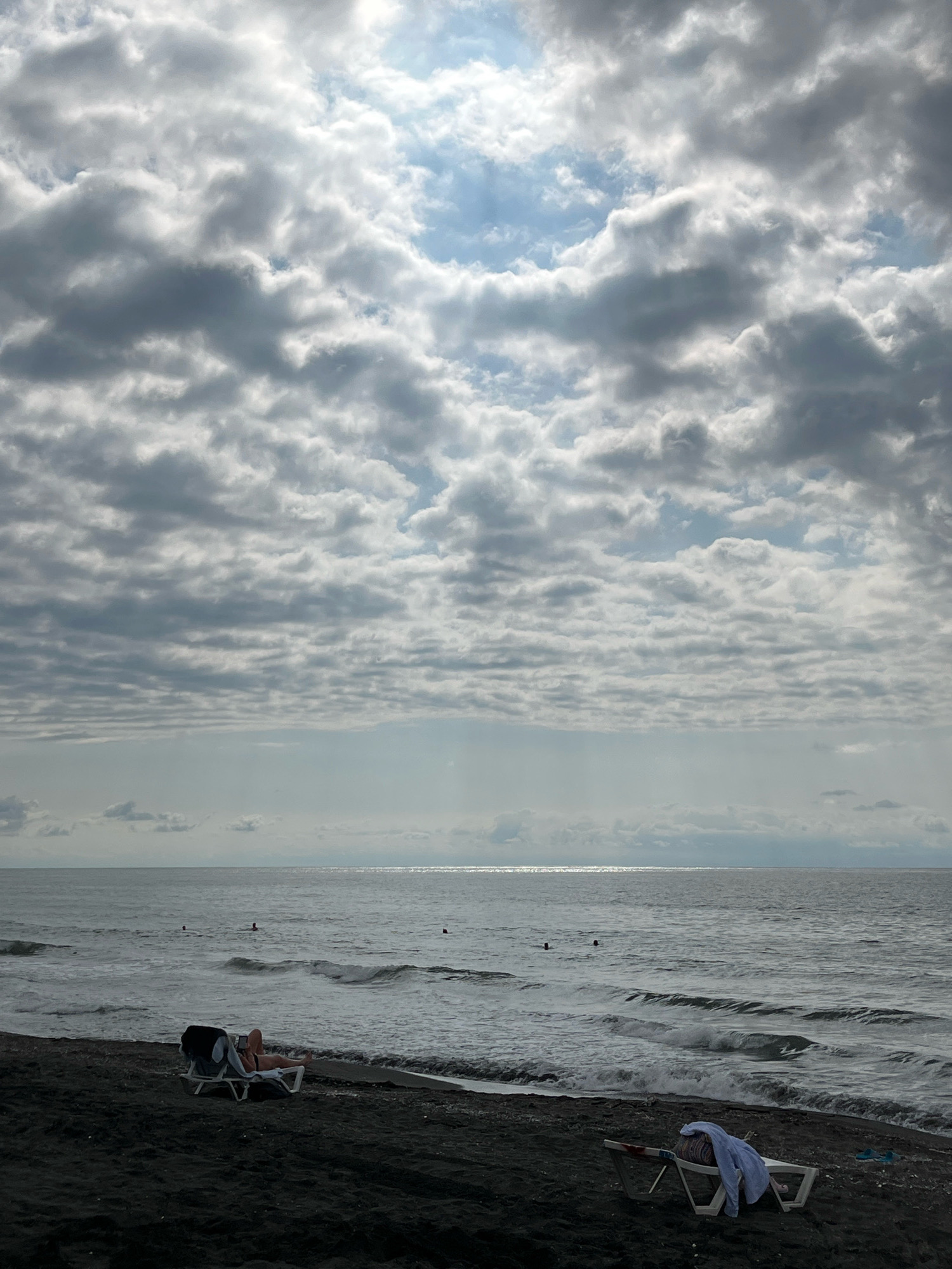 The image depicts a cloudy day at a beach in Ureki, Georgia. In the foreground, a dark sand beach is visible with two lounge chairs. One lounge chair on the left has a person lying on it, wearing a bikini. The other lounge chair on the right is empty and has a white shirt and blue flip-flops draped over it. The sea is calm with gentle waves rolling onto the shore. Several people are seen swimming or wading in the water in the middle ground. The sky is filled with dramatic, layered clouds, with patches of sunlight breaking through. The horizon line is clearly defined between the sea and the sky, with a subtle haze or mist extending across the water. The overall atmosphere is overcast, suggesting a cool, possibly humid day.