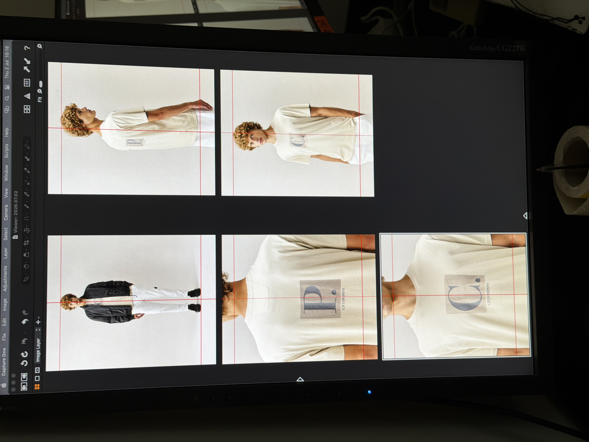 The image displays a computer monitor, likely a professional photography or design workstation, showing a layout of four different images of a male model. The model is wearing casual clothing, including a bomber jacket and white pants in the top left image, and a light-colored t-shirt with a graphic logo in the other three. The images appear to be product shots, possibly for an apparel brand. The monitor's interface, featuring menu bars and tool icons, is also visible. Red guide lines, typical for composition and cropping in image editing software, are overlaid on each of the model's images. The environment surrounding the monitor is dimly lit and appears to be an office or studio space, with the edge of another monitor and some desk clutter, including a roll of tape and a pen, visible. The specific location in Milan, Italy, cannot be determined from the provided image.