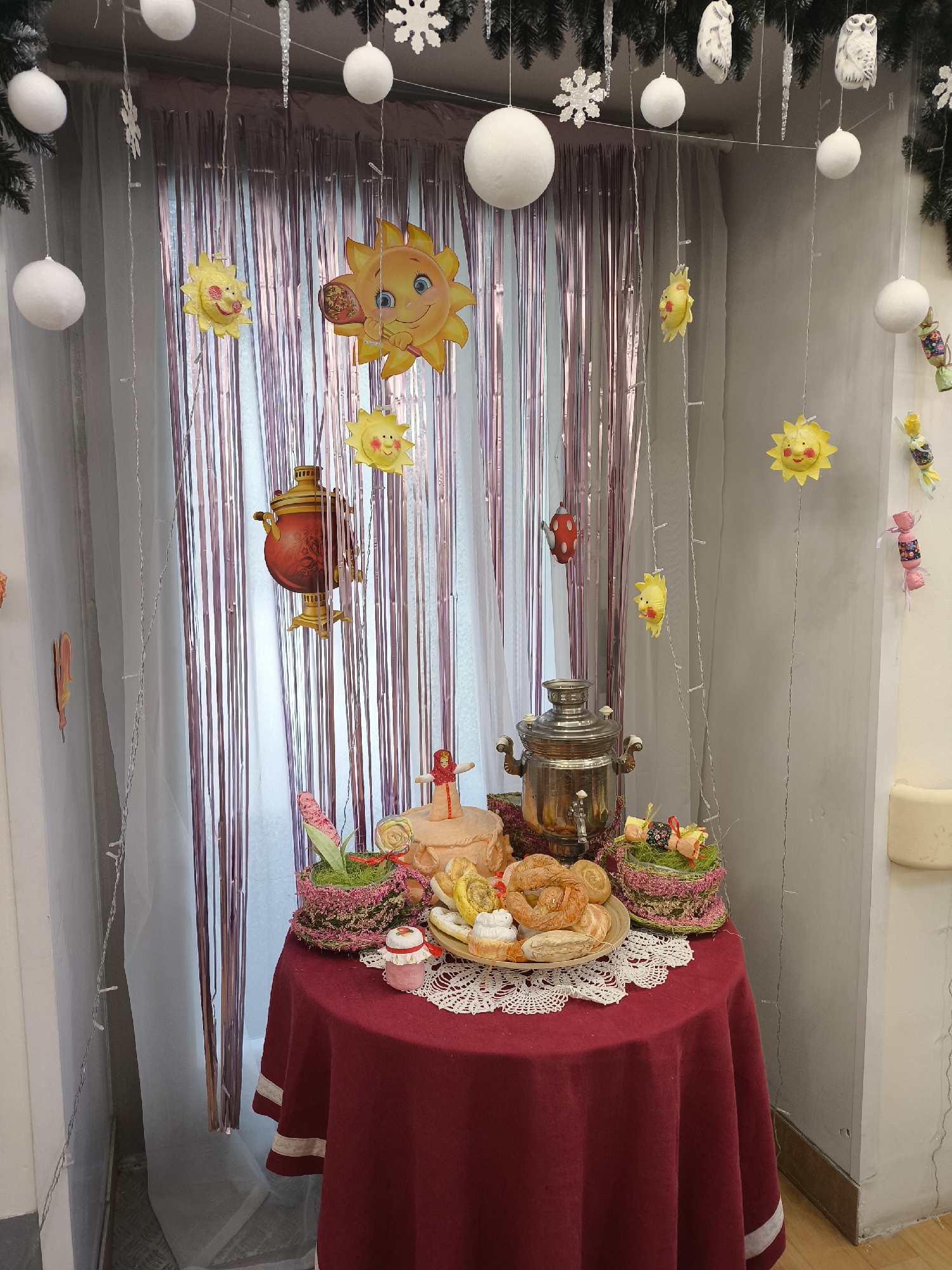 A festive table adorned with traditional Russian items stands in front of a window in what appears to be a room in Moscow, Russia. The window is decorated with strands of garland containing snowflakes and white spheres, as well as cut-out images of smiling suns and candies. A red tablecloth with a white border covers a round table upon which sits a silver samovar and a painted tea pot. Various foods including bread and small cakes are arranged on a plate and in baskets. Decorative items, such as small Easter eggs and flower arrangements are also present on the table. The room is sparsely decorated with white walls, adding to the focus on the table arrangement.