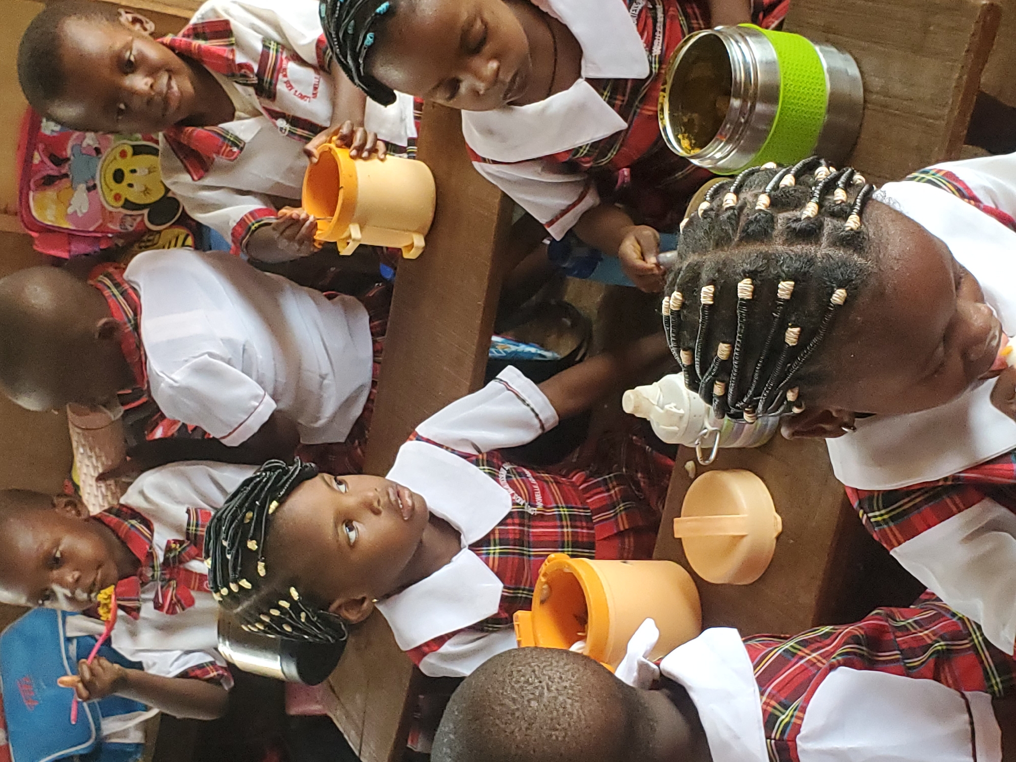 In Nsugbe, Nigeria, a group of school children, presumably in elementary school, are eating their lunches. They are sitting at wooden desks. The children are wearing school uniforms that are predominantly white with a red and green plaid pattern. Some of the children are using plastic utensils to eat from yellow and green containers. One child has a blue backpack with a cartoon character on it. Their hair is styled with braids.