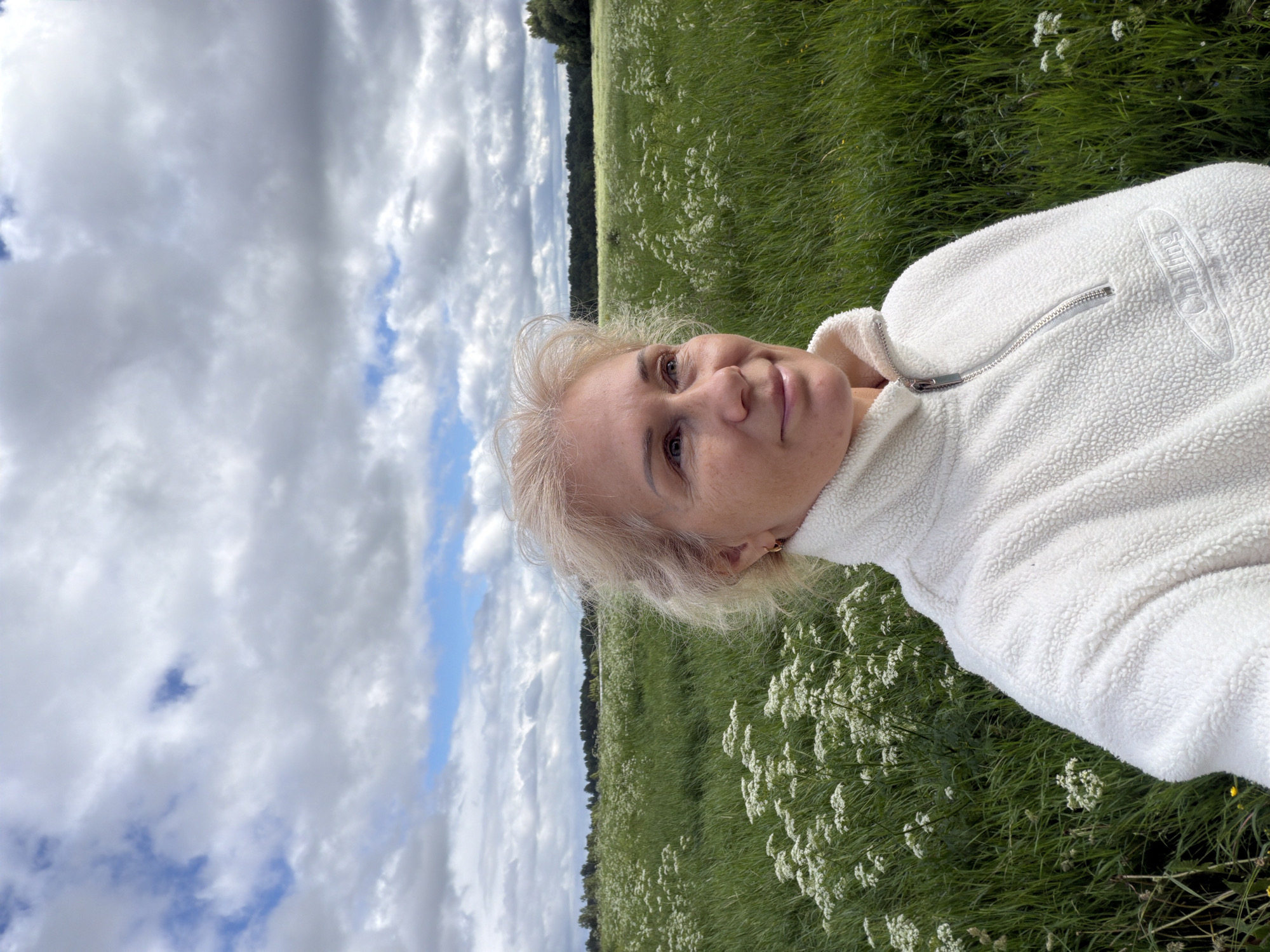 A medium-shot selfie captures a middle-aged Caucasian woman with blonde, wavy hair and blue eyes. She is wearing a cream-colored, fuzzy fleece half-zip sweater. Her head is turned slightly to the right, and she is looking upwards and to the right with a subtle smile. The background is a vast, green field dotted with white wildflowers, extending to a dark tree line under a cloudy sky. The scene appears to be outdoors in a rural or natural setting. The location is Steklyannyi, Russia.