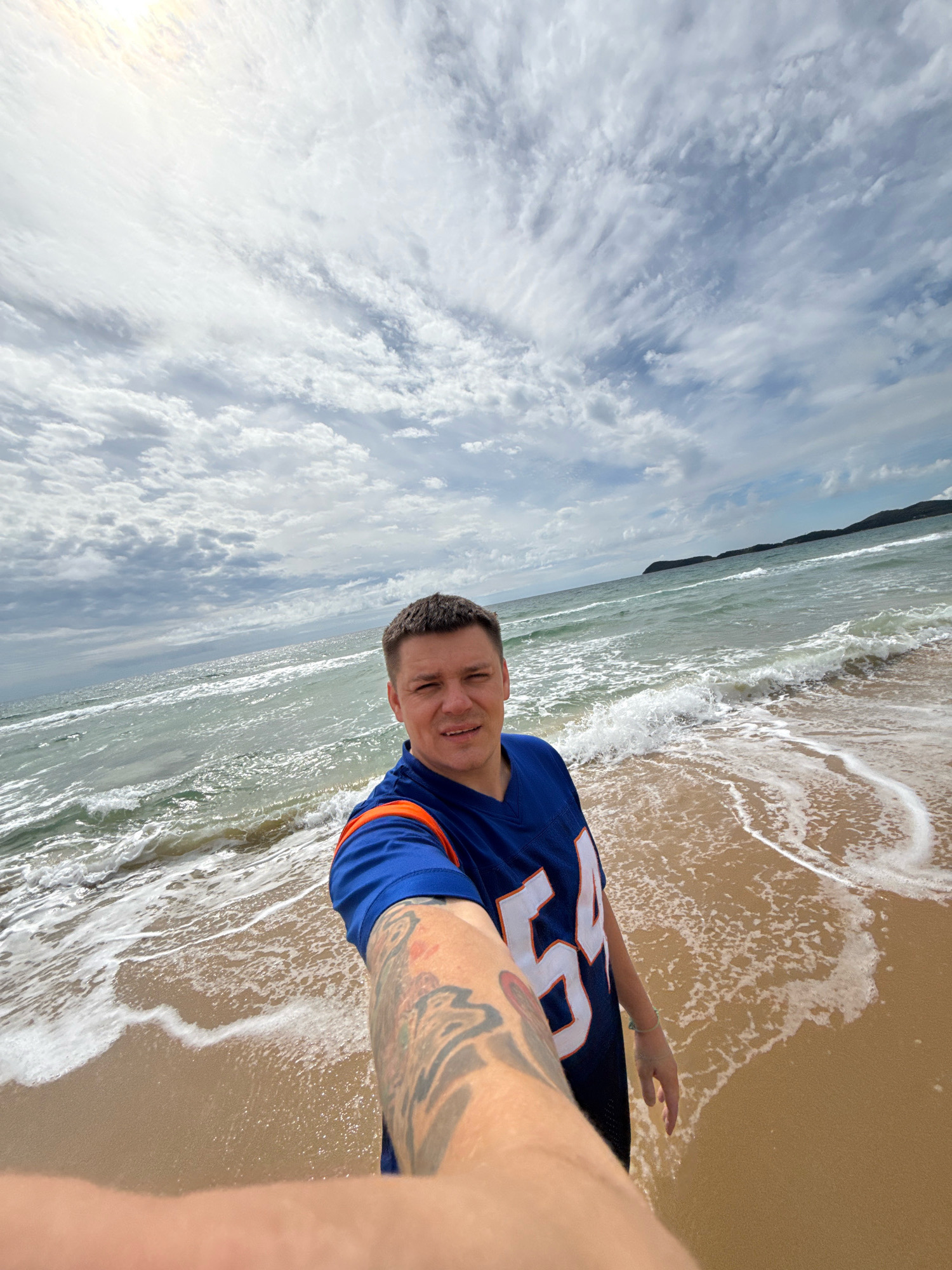 A man in a blue football jersey with the number "54" on it takes a selfie at the beach. He is standing on wet sand with waves gently breaking on the shore. The sky is filled with white, wispy clouds. In the background, the ocean stretches to the horizon, with a faint outline of hills on the right side. The man's right arm, which is tattooed, is extended towards the camera, holding it. The overall impression is a casual moment captured during a beach outing. The specific location in Anna, Russia, is not discernible from the image itself.