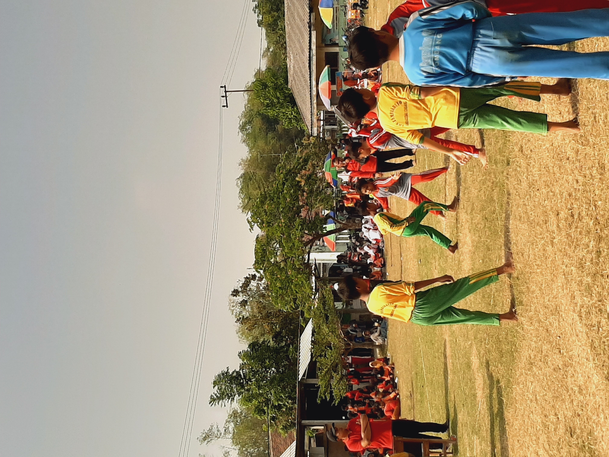 An outdoor daytime event is taking place on a grassy field in Sumberjo, Indonesia. In the foreground, several children are dressed in sportswear. Two boys in yellow shirts and green pants are the most prominent figures. One boy in a blue tracksuit with white writing on the back stands facing away from the camera. Another boy in a yellow shirt and green pants with yellow stripes on the side is also visible. Several other children in similar athletic attire are scattered across the field, some appearing to be participating in an activity, while others are observing.

In the midground, there are more children and adults, some seated and others standing. Several colorful umbrellas and canopies are set up, suggesting a gathering or event. Buildings, possibly a school or community center, are visible in the background, along with trees and utility poles. The overall scene depicts a lively, organized event, likely a school sports day or similar community gathering. The lighting suggests it is a sunny day.