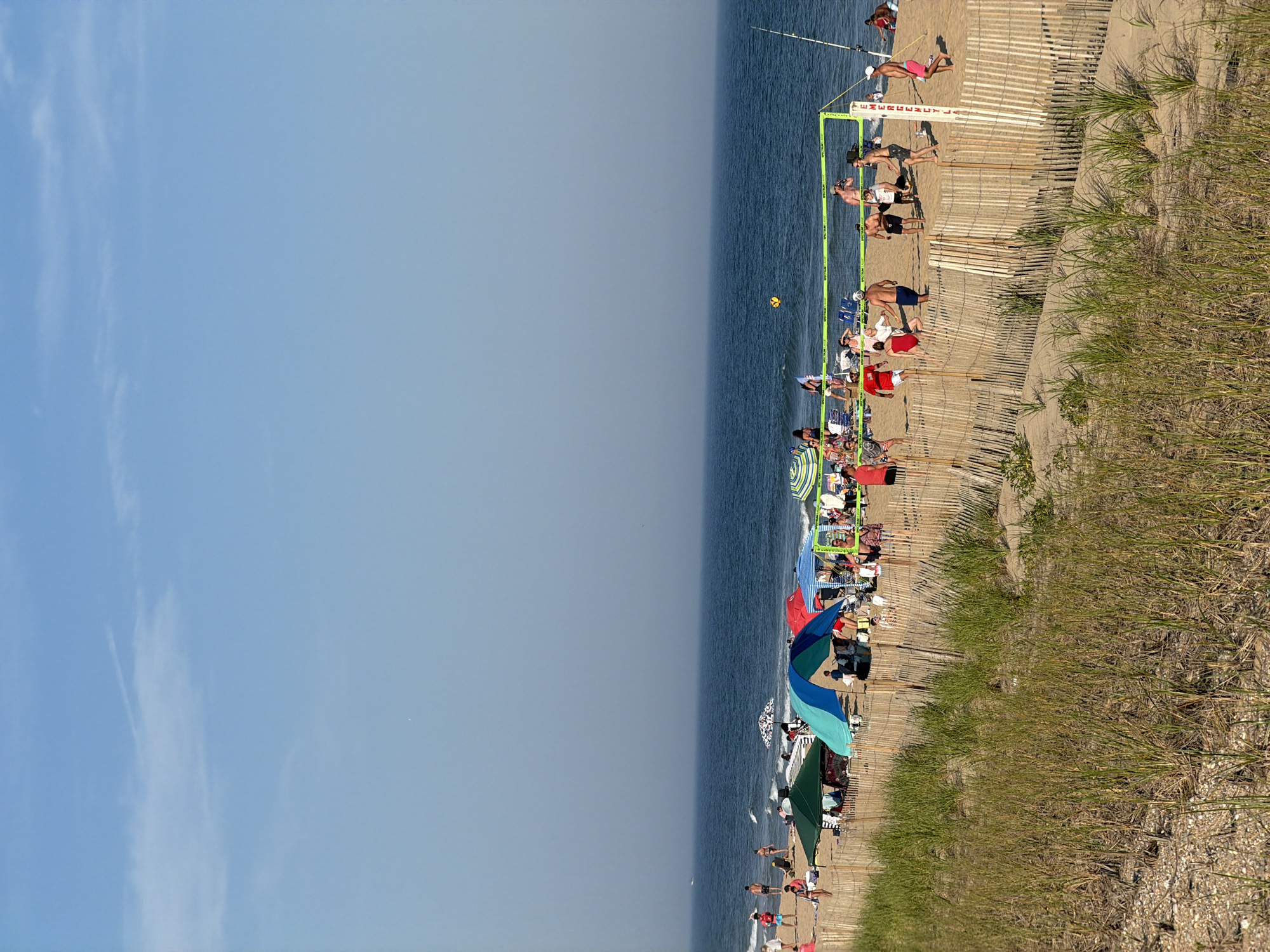 In Montauk, United States of America, a sunny day unfolds on a crowded beach. The ocean, a deep blue, stretches to the horizon under a clear sky with a few wispy clouds. The sandy shore is dotted with numerous beachgoers enjoying various activities. A group of people is engaged in a lively game of beach volleyball, with players positioned on either side of a bright green net. Nearby, beach umbrellas and tents provide shade for sunbathers. A wooden fence runs along the edge of the beach, separating it from a sandy dune covered in tall, dry grass. The scene captures a typical summer day at the beach, full of recreation and relaxation.