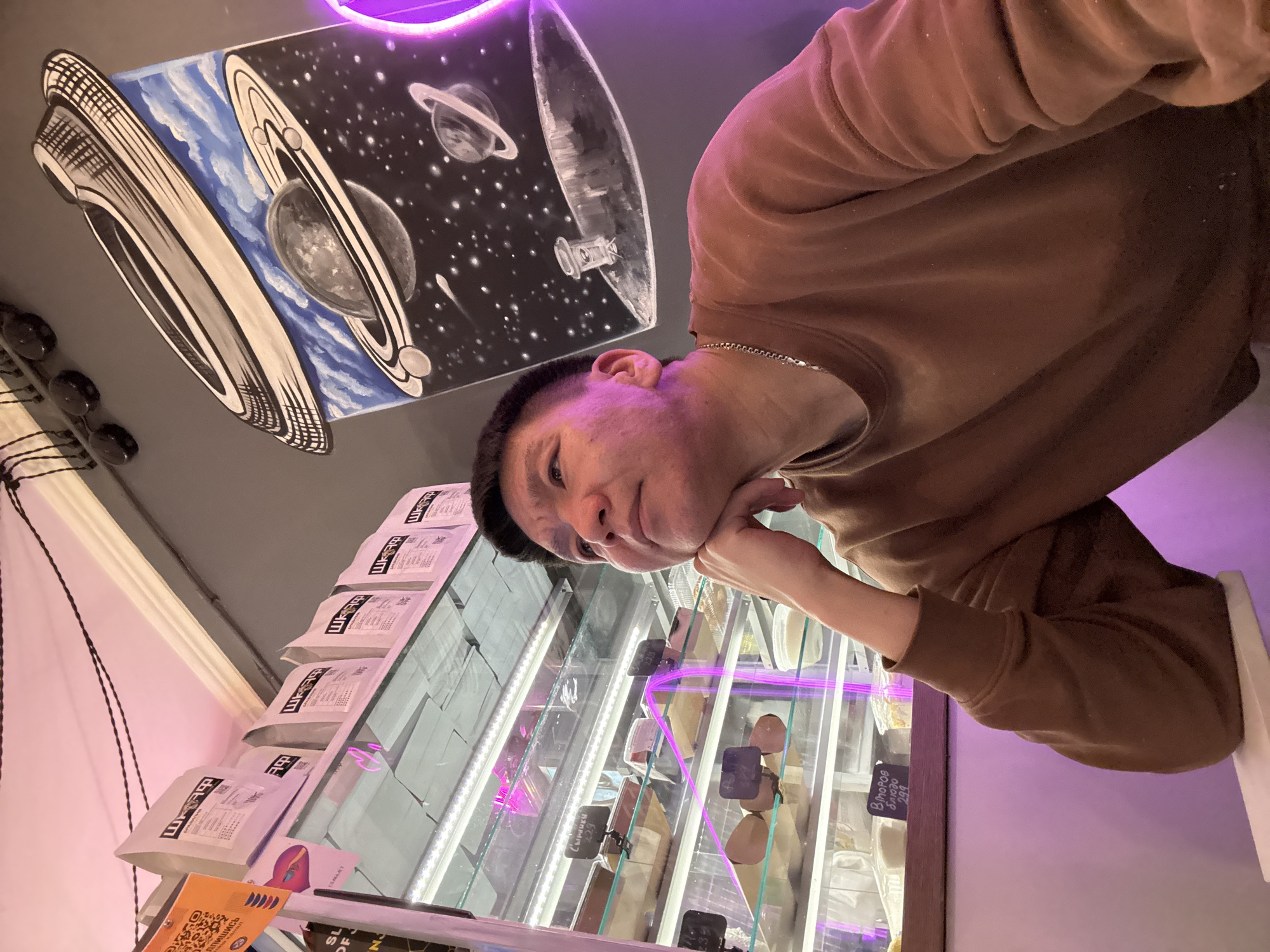 A man is pictured in a cafe in Moscow, Russia. He is wearing a brown sweatshirt and a silver chain. He is leaning his chin on his hand and looking upwards. The background shows shelves of coffee bags and a display case filled with food items. A painting of a space scene with planets hangs on the wall above the man. The cafe is illuminated by pink and purple neon lights.