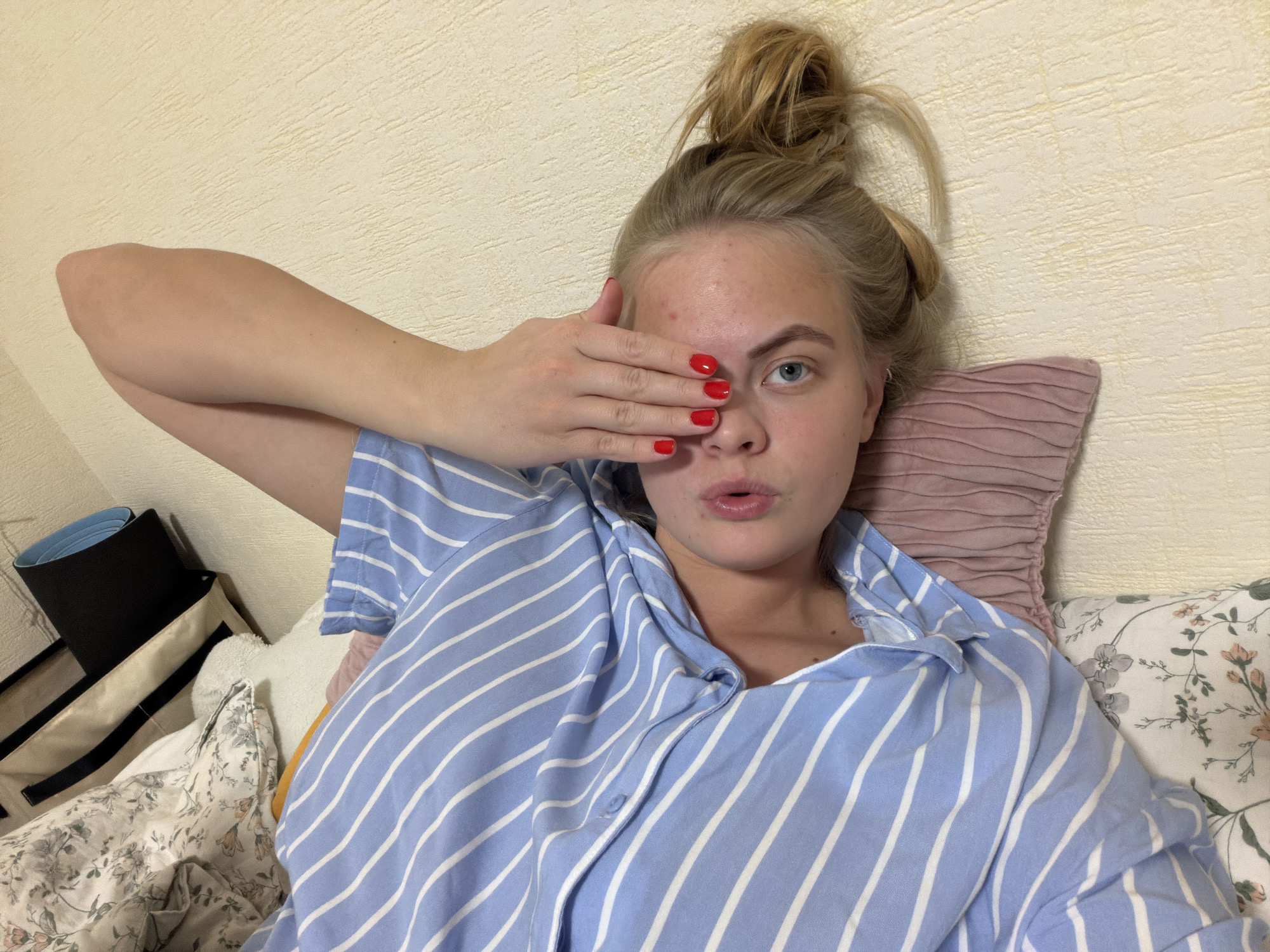 A young woman is lying in bed, covering one eye with her hand. She is fair-skinned with blonde hair pulled up in a messy bun. She is wearing a blue and white striped shirt. Her fingernails are painted red. She is lying on a bed with various pillows behind her. The wall behind the bed is textured and appears to be cream-colored. A black yoga mat and a beige bag with black handles are visible on the left side of the frame. The location is likely an interior bedroom space, and there is no evidence to suggest it is in Izhevsk, Russia.