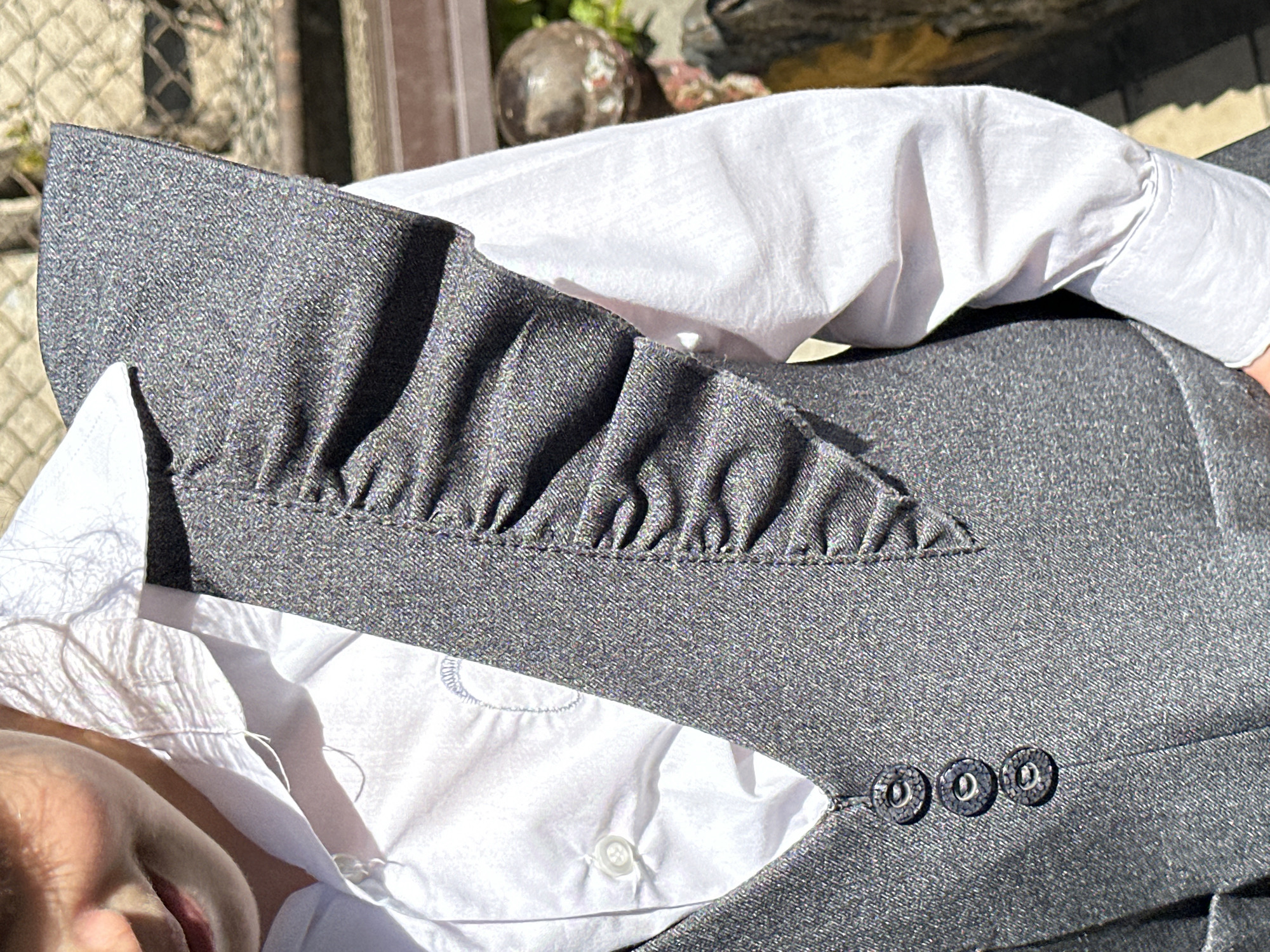 This close-up, angled photograph, taken outdoors in Agarak, Armenia, captures the upper torso of a person, likely a child, dressed in formal attire under bright, direct sunlight. Strong shadows fall across the white shirt, indicating a sunny daytime setting.
The subject is wearing a grey suit jacket over a white collared shirt. The jacket is made of a finely textured, possibly tweed or woven fabric, which appears to have subtle iridescent flecks reflecting the intense sunlight. A distinct design element is the series of prominent ruffles or fine pleats along the inner edge of the jacket's lapel. Three dark, round buttons with metallic-looking rims are visible on the jacket's front, arranged vertically.
Beneath the jacket, a crisp white collared shirt is worn, with a single white button visible on its placket. A subtle, light grey embroidered detail is visible on the shirt's opening, peeking out from under the jacket. A small portion of the person's face, including a cheek and mouth, is partially visible in the bottom-left corner of the frame. The background is blurred, showing hints of a metal fence or grating, and some decorative garden elements like a spherical object and foliage, consistent with an outdoor environment. No specific activity is discernible beyond the person being dressed. There is no visible text in the image.