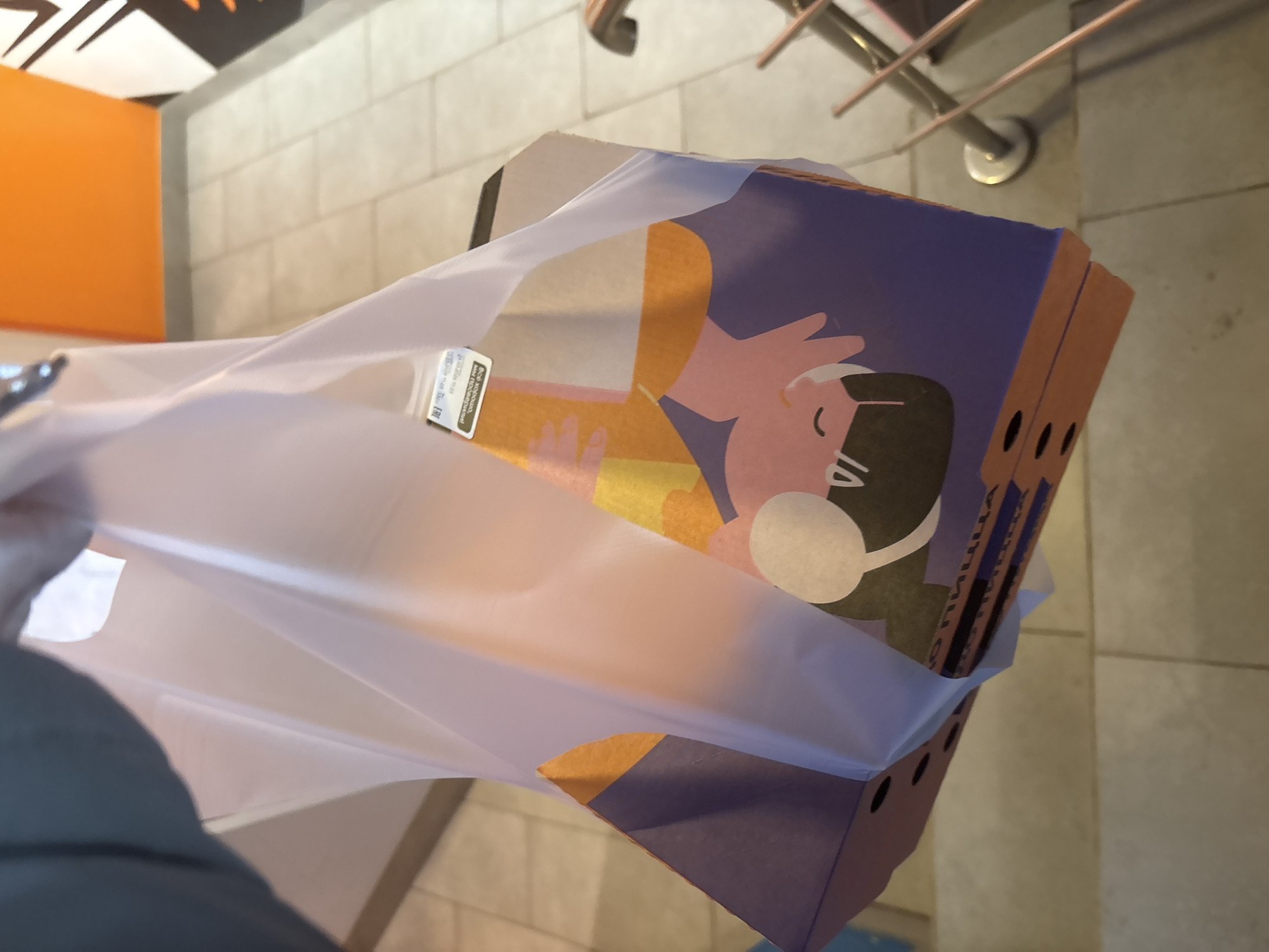 The image shows a purple cardboard box in a clear plastic bag. The box has a cartoon illustration of a person with headphones on. The background shows a tiled floor and part of a wall with an orange panel. Based on the limited information, it is difficult to definitively confirm the location in Votkinsk, Russia.