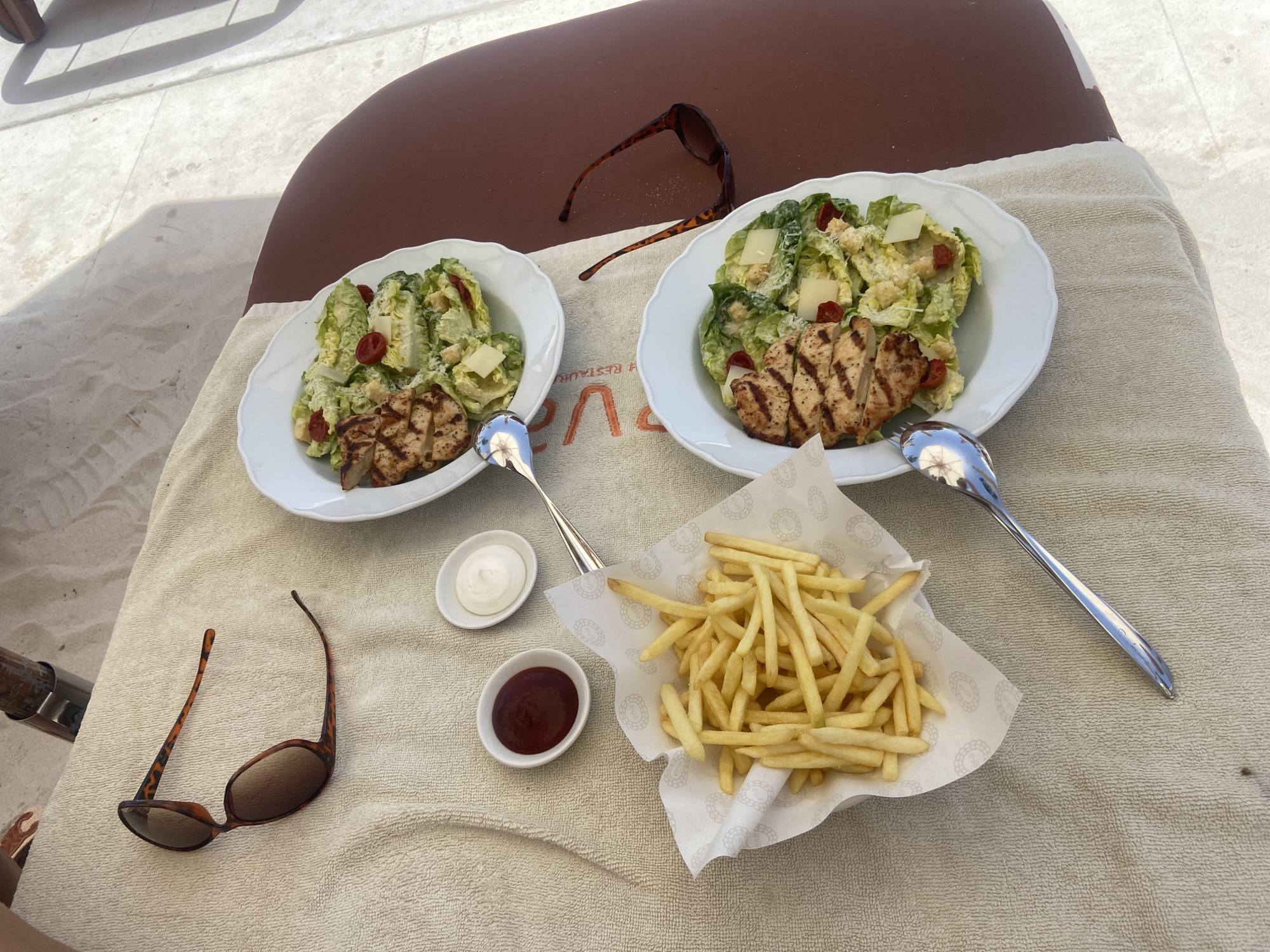 The image captures an outdoor dining scene in Dubai, United Arab Emirates, set upon a brown chaise lounge draped with a beige towel. The setup suggests a relaxed, possibly beachside or poolside setting. Two white scalloped-edged plates each contain a Caesar salad with grilled chicken. Accompanying these are two silver spoons, small white dishes filled with white and red sauces (likely mayonnaise and ketchup), and a portion of french fries served in a patterned paper container. Two pairs of tortoise-shell sunglasses are placed on the lounge, indicating a sunny environment. The presence of food and dining utensils points to a meal being consumed or recently finished in a casual outdoor setting. The background is blurred, but the neutral tones suggest a sandy or paved area, typical of Dubai's beach or resort environments.
