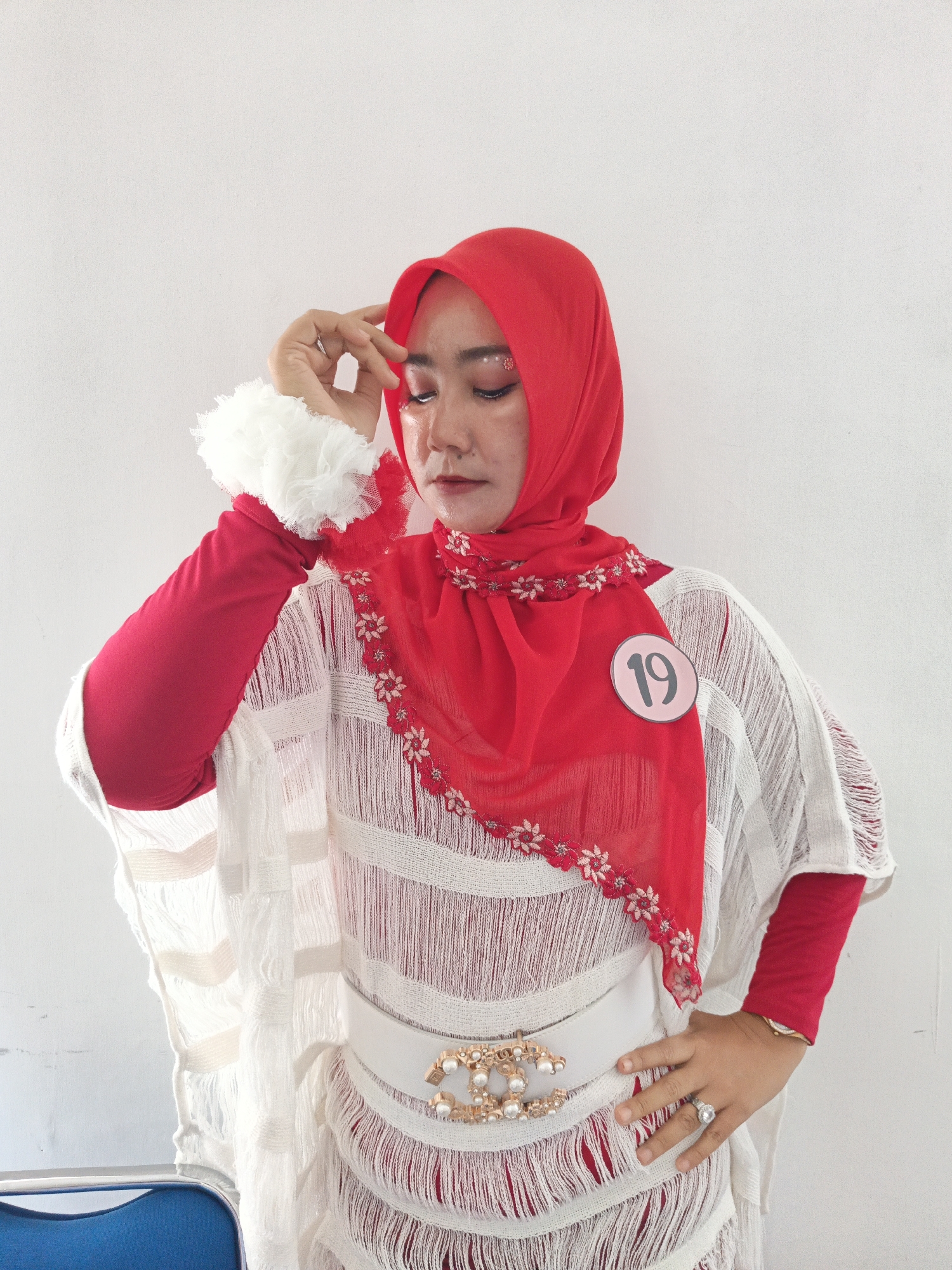 A young woman is posing in front of a white wall, likely in Klaten, Indonesia. She is wearing a red hijab and a white, fringed outer garment over a red long-sleeved shirt. The white garment has horizontal stripes of varying thickness. A white belt with a large, ornate gold and pearl buckle cinches her waist. She has a pink circular tag with the number "19" on her right side, attached to her red hijab. Her makeup includes glitter on her eyelids. Her left hand is raised to her forehead, and she is wearing rings on her fingers. A blue chair is partially visible in the lower left corner of the frame. The overall lighting suggests an indoor setting with a bright light source.