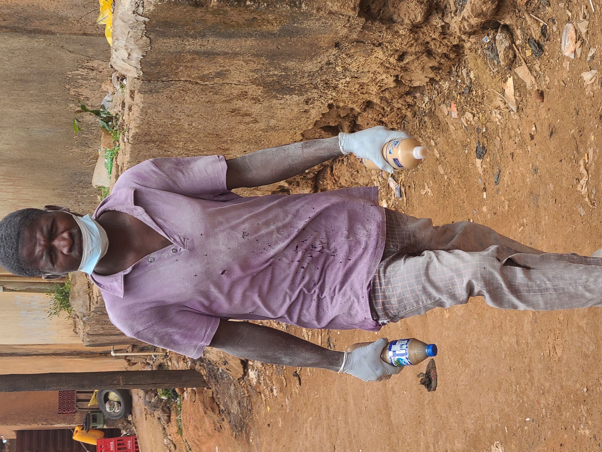 A man wearing a purple, stained polo shirt, plaid pants, and a blue surgical mask is holding two bottles of liquid with both hands, each covered in white gloves. The man is positioned outdoors in an area containing dirt and debris, possibly near a building. The location is Jos, Nigeria.