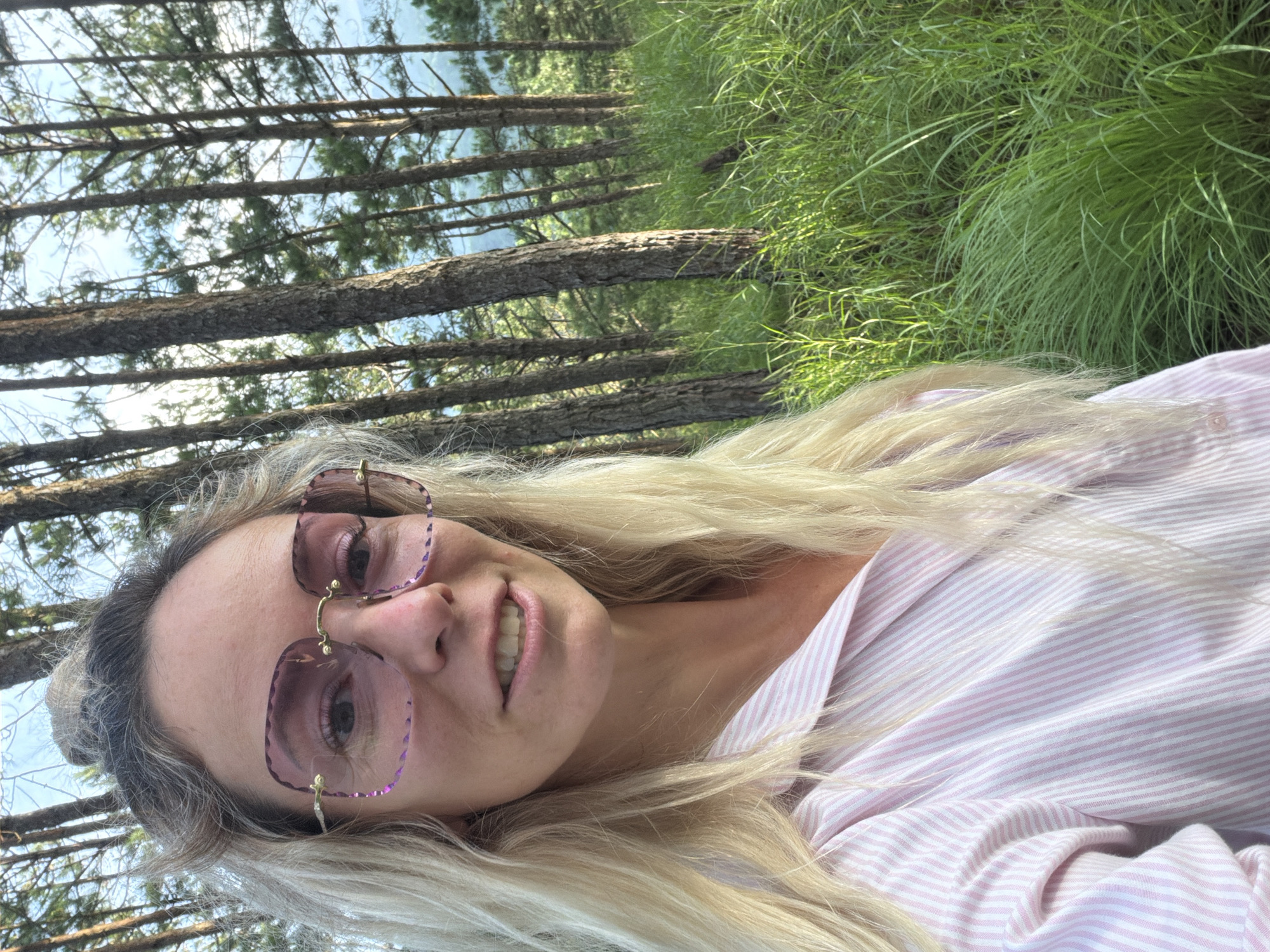 A woman with blonde hair, wearing oversized pink-tinted square glasses and a pink and white striped button-up shirt, stands in a forest with tall pine trees and lush green grass. The background shows more trees and a hint of the sky. The woman is looking towards the camera and smiling. The location appears to be Xuan Truong - Da Lat, Vietnam.