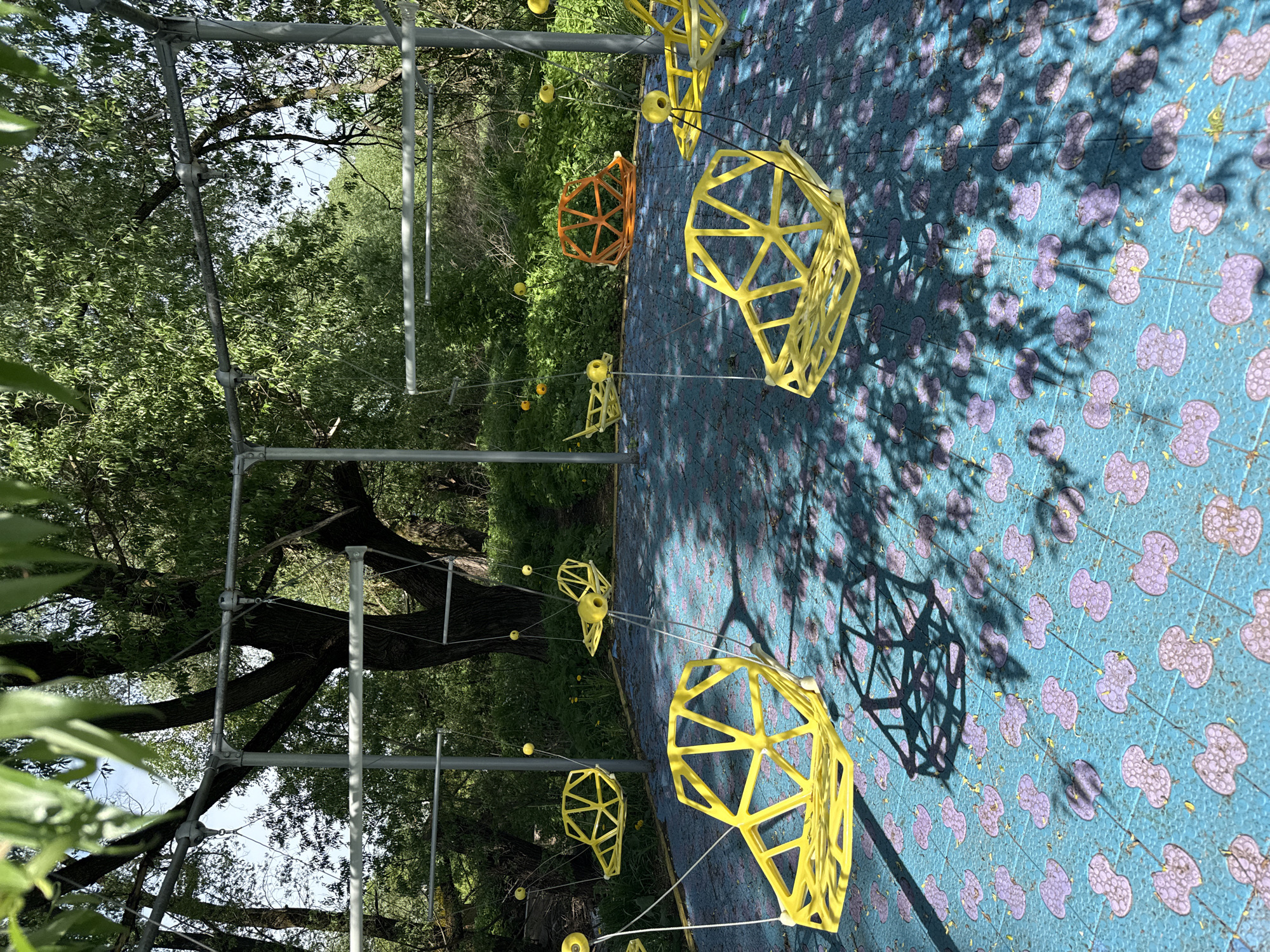 A partially inverted eye-level outdoor shot taken in Синьково, Russia, presents a geometrically themed playground or park installation against a natural backdrop of trees and vegetation. The image is rotated counterclockwise, so the intended “ground” appears on the right.
In the foreground and central area, several geometric structures made of yellow and orange materials are suspended or placed on a blue surface patterned with purple clover-like shapes. These structures resemble hollow polyhedrons or partial geodesic domes, some casting stark shadows on the patterned ground. They are connected with thin, white strings to silver metal poles. The geometric shapes appear to be part of a playground installation.
In the background, a green landscape is visible, including trees, plants, and hints of other park elements. Metal poles support the structures. The light suggests it is daytime with clear skies.