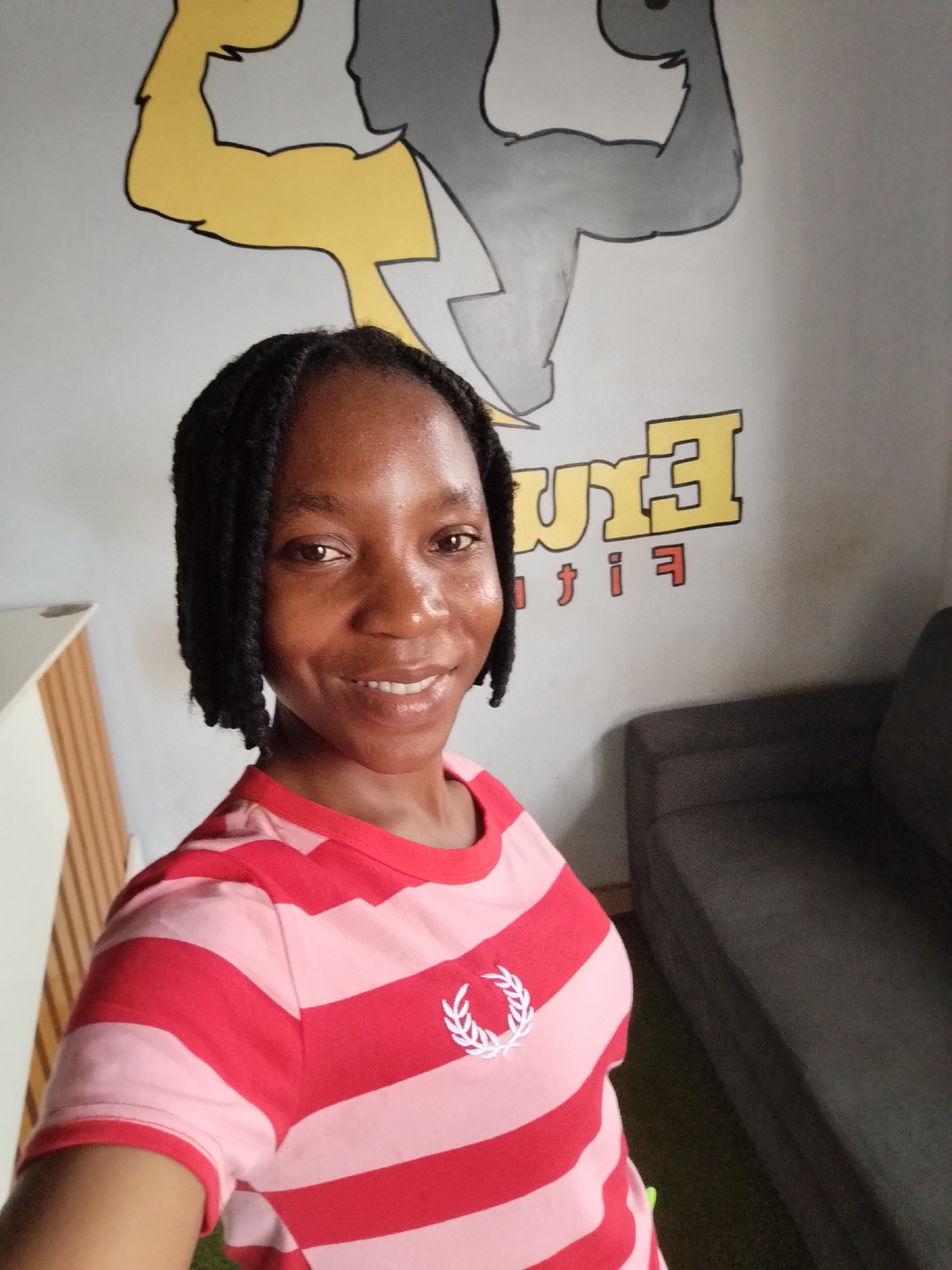 A young Black woman with braided hair smiles at the camera in an indoor setting. She is wearing a pink and red striped t-shirt with a white laurel wreath emblem on the left chest. The background features a mural of two muscular silhouettes, one yellow and one gray, flexing their biceps. Below the silhouettes, text reads "ERIE FIT" in yellow and red lettering. To the left of the woman is a white cabinet with vertical wooden slats, and to her right is a dark gray sofa. The overall environment suggests a gym or fitness studio in Uselu, Nigeria.