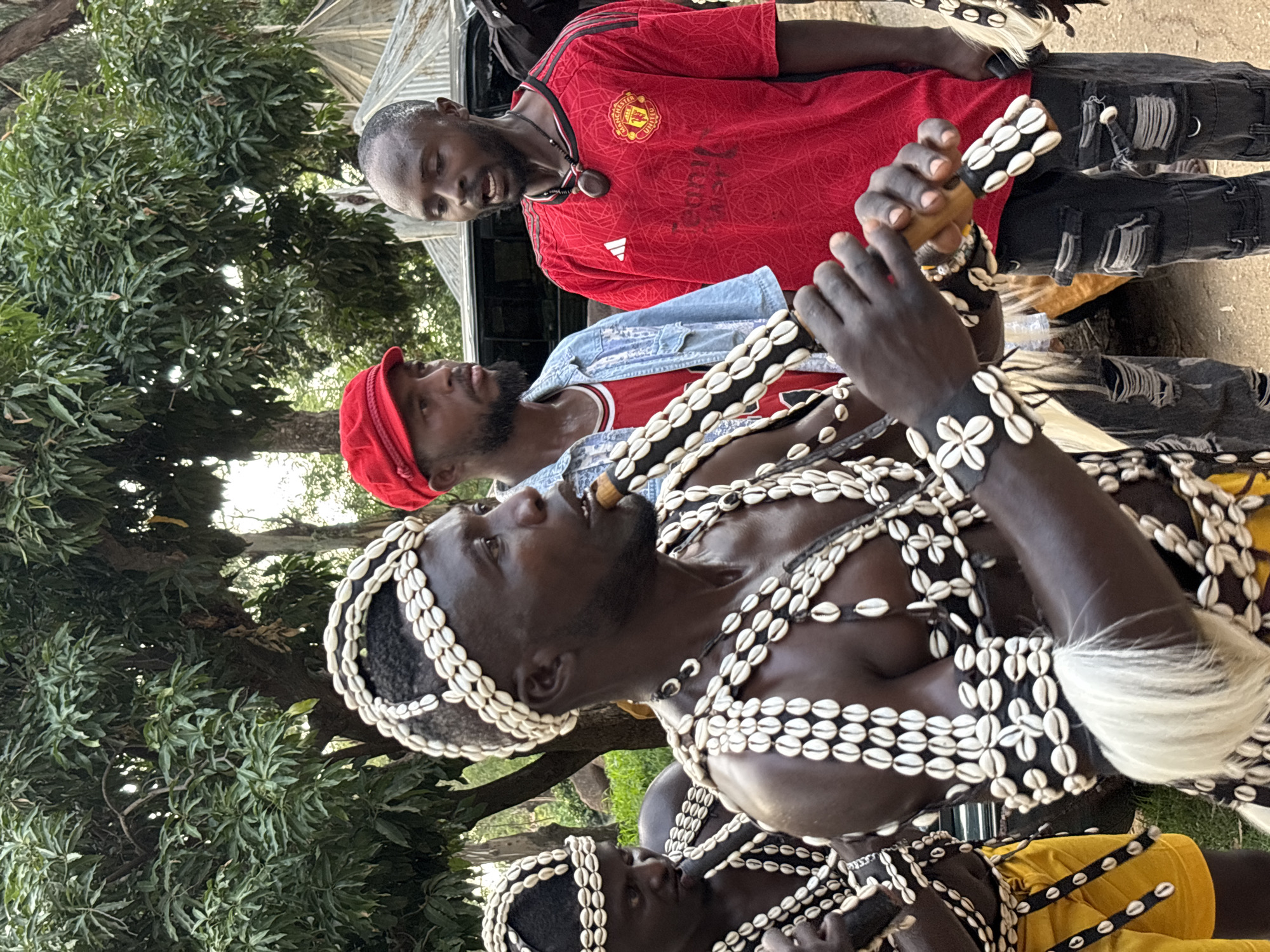 In the city of Jos, Nigeria, a group of men are gathered outdoors, with lush green trees forming the backdrop. The primary subject, a man in the foreground, is adorned in traditional attire featuring intricate shell decorations on his head and chest. He is playing a wooden flute-like instrument, his lips pressed against it. To his left, another man, also in similar shell-decorated attire, is partially visible, also appearing to play an instrument. To the right of the foreground subject, a man in a red Manchester United jersey with a wooden pendant around his neck is looking towards the foreground. Behind him, another man wearing a red cap and a denim jacket over a red shirt is also present. The setting appears to be a cultural gathering or performance, with a car visible in the background. The overall atmosphere suggests a vibrant community event.